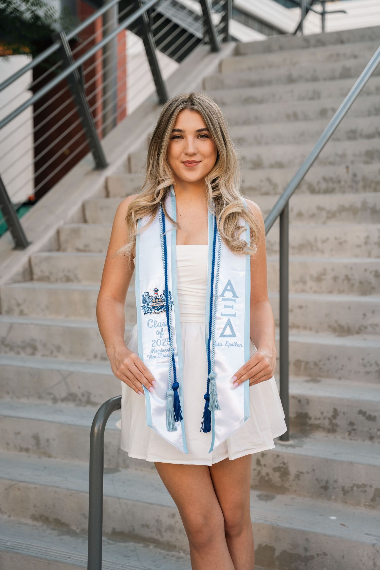 Graduate wearing white academic stole poses on outdoor campus steps.
