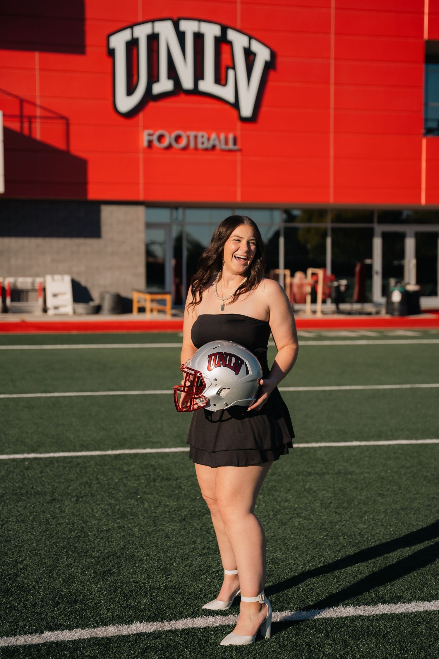 Graduate in black dress smiles while holding UNLV football helmet on field.