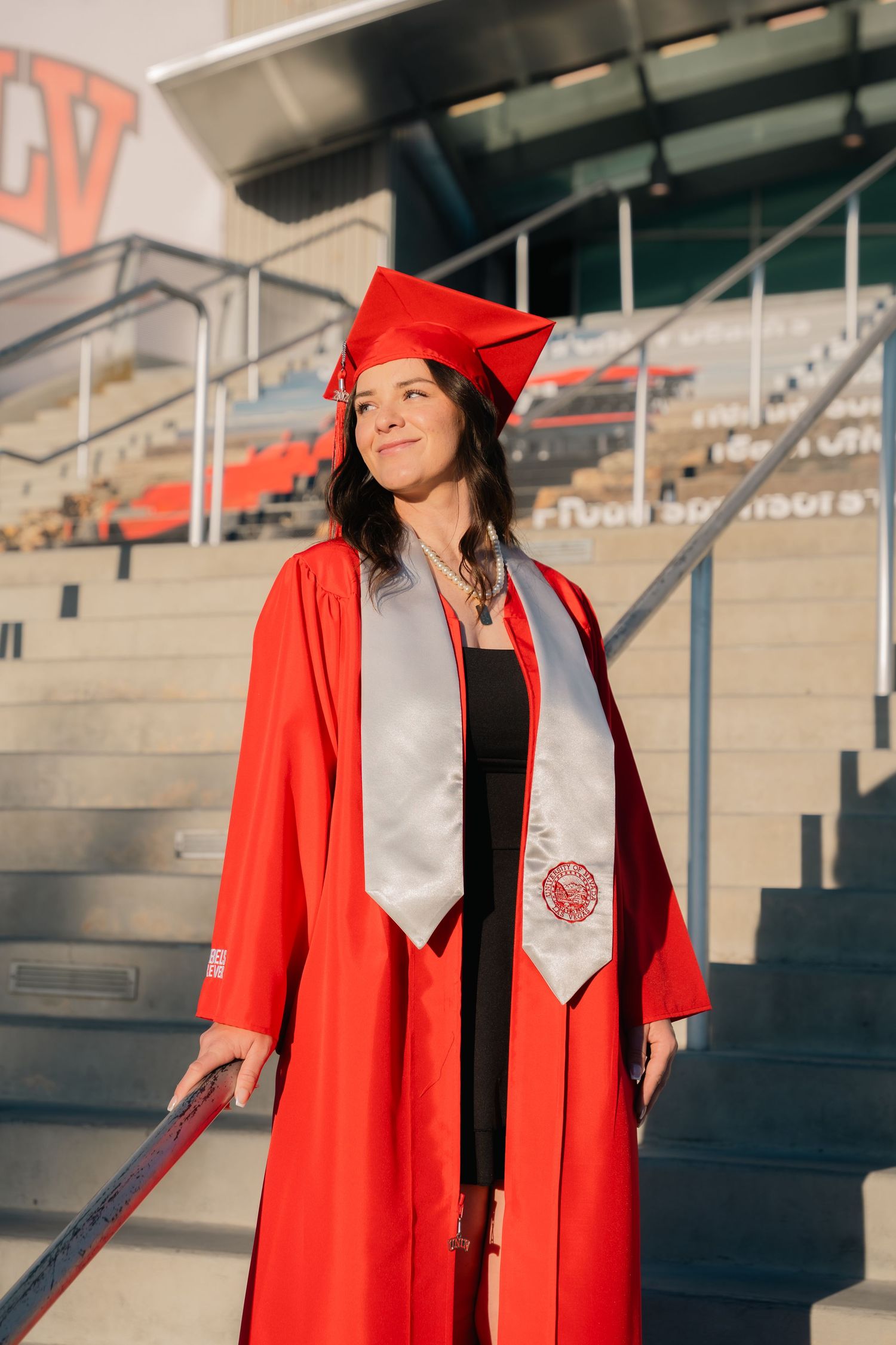 Graduate in red cap and gown poses near stadium steps.