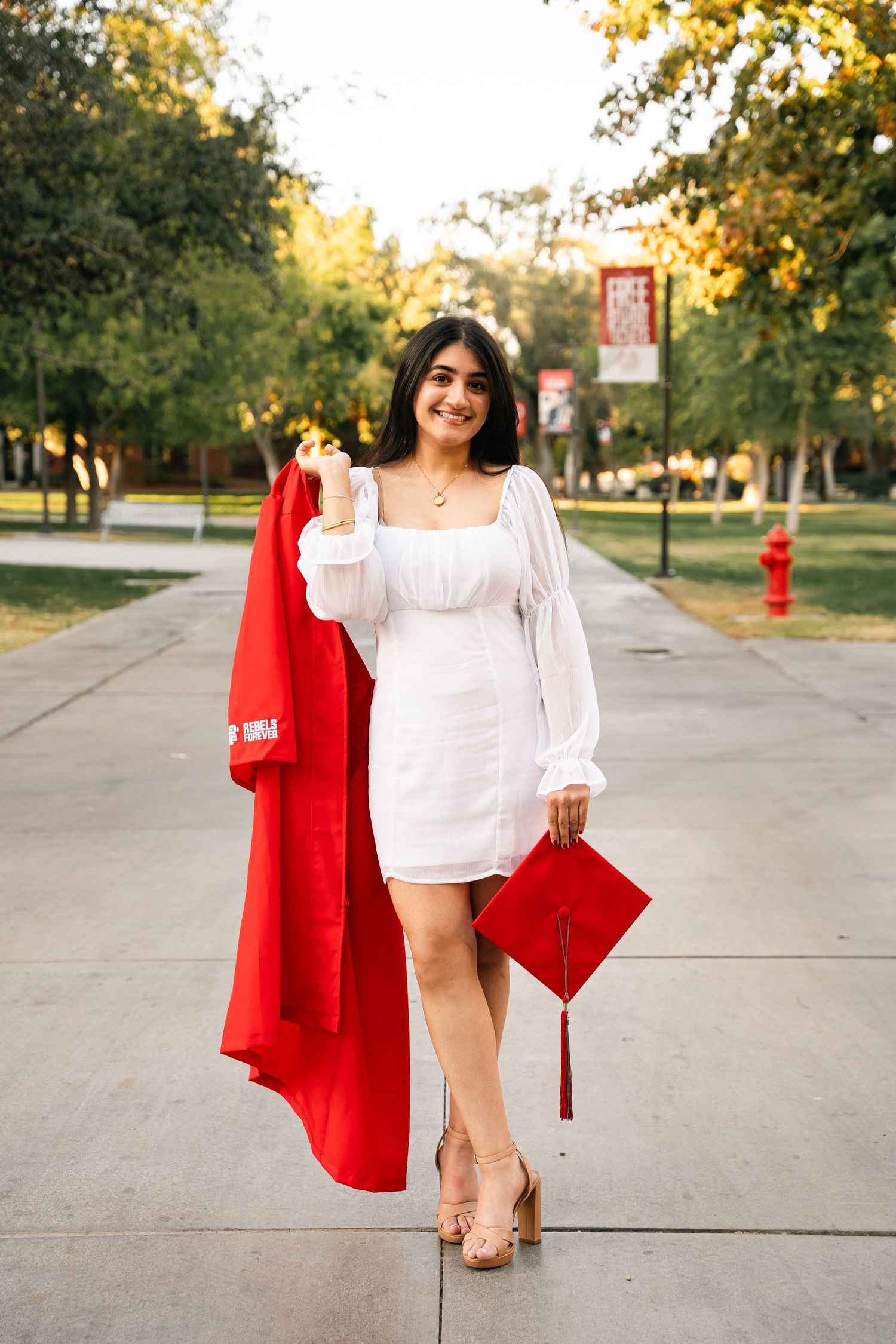 Graduate in white dress carrying red graduation gown and cap on university walkway.