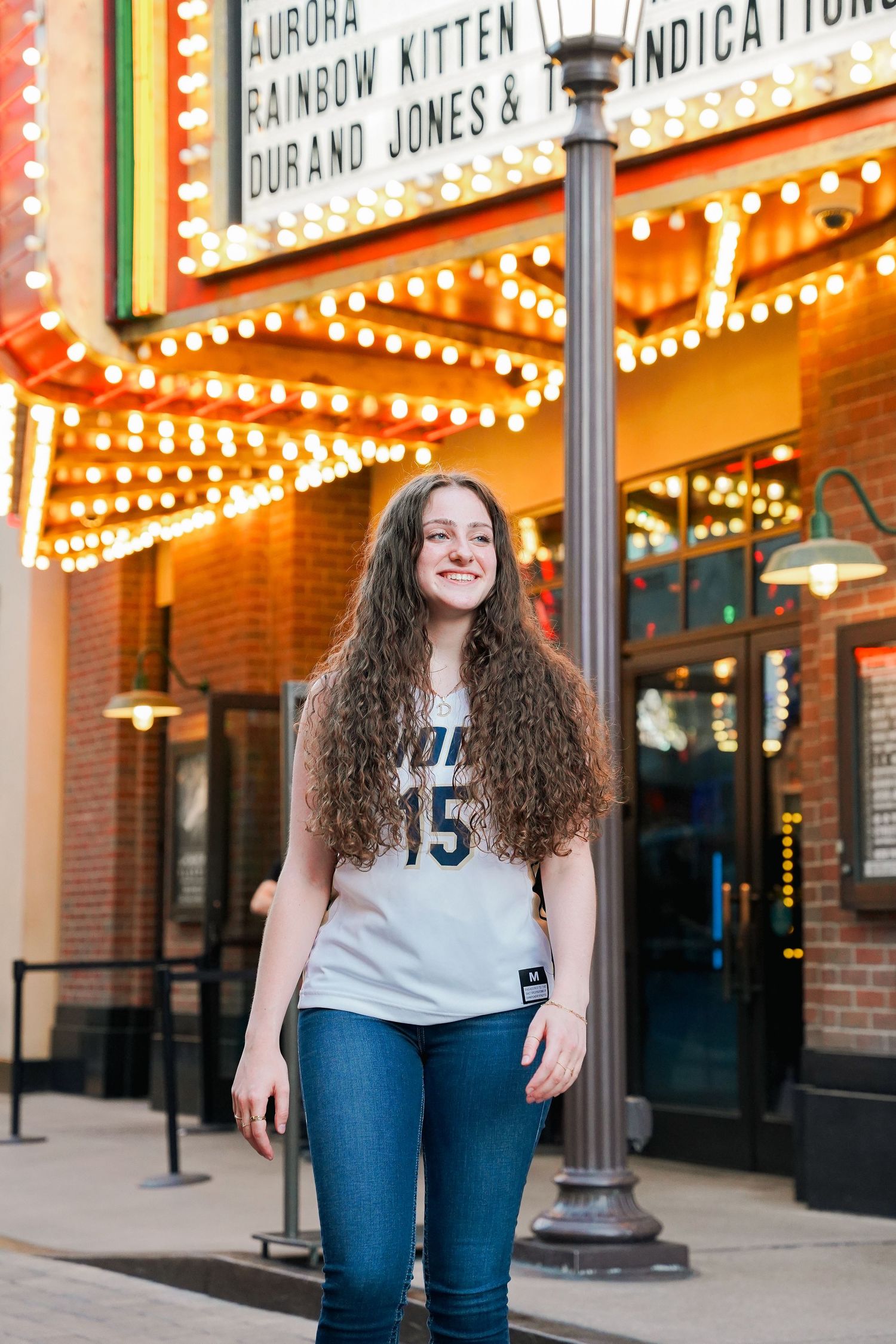 Person with long curly hair wearing Lions #15 jersey poses in front of illuminated theater marquee.