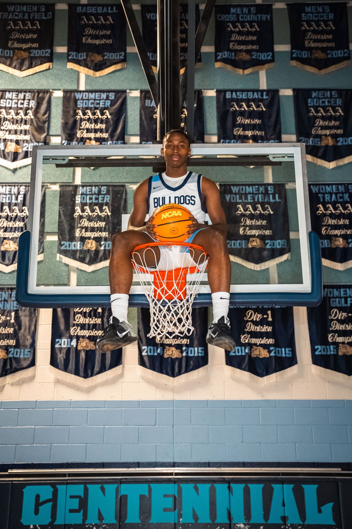Basketball player sits on backboard holding ball with championship banners behind.