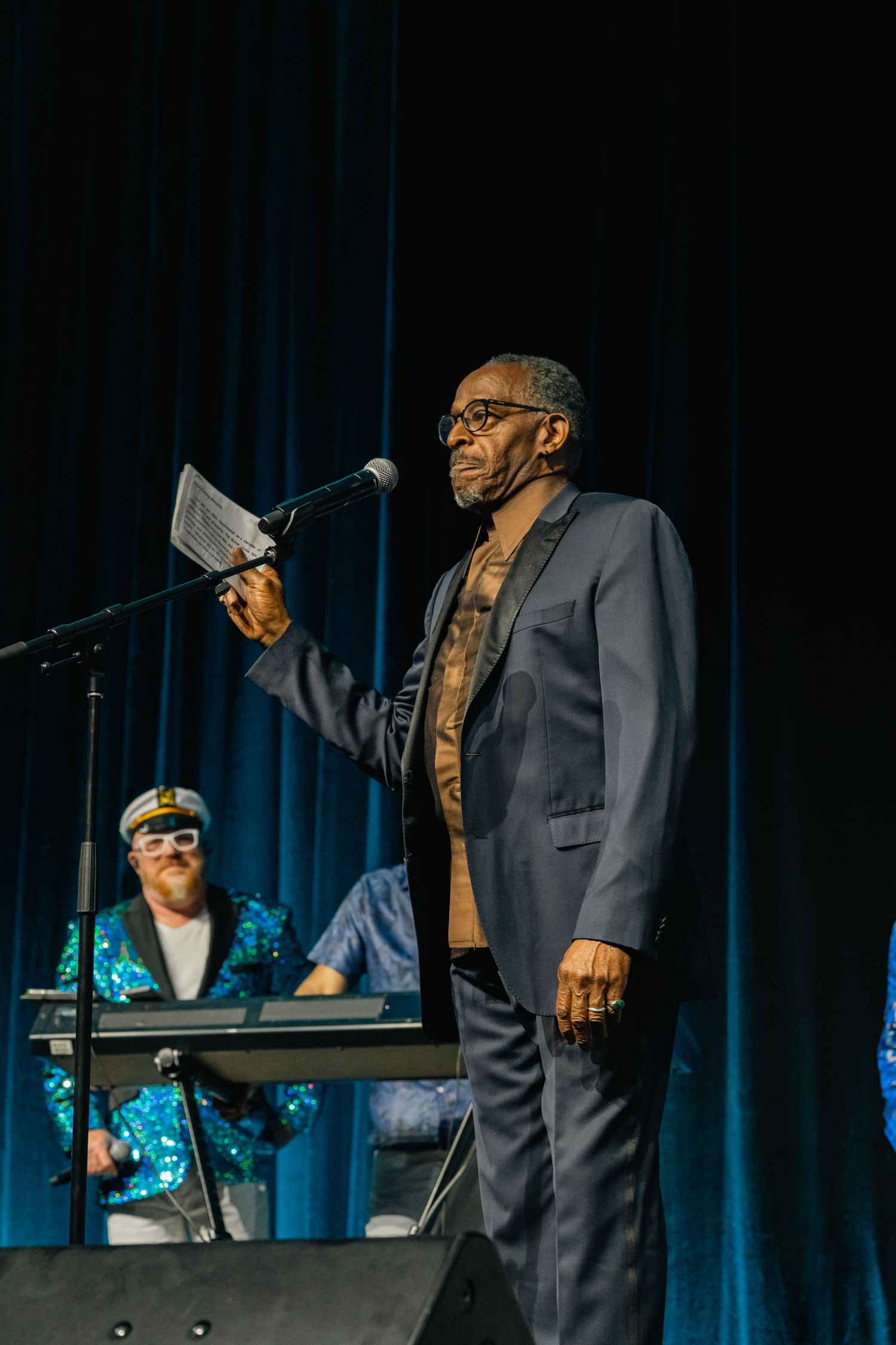 Performer in suit jacket speaking into microphone on stage with keyboard player in background.