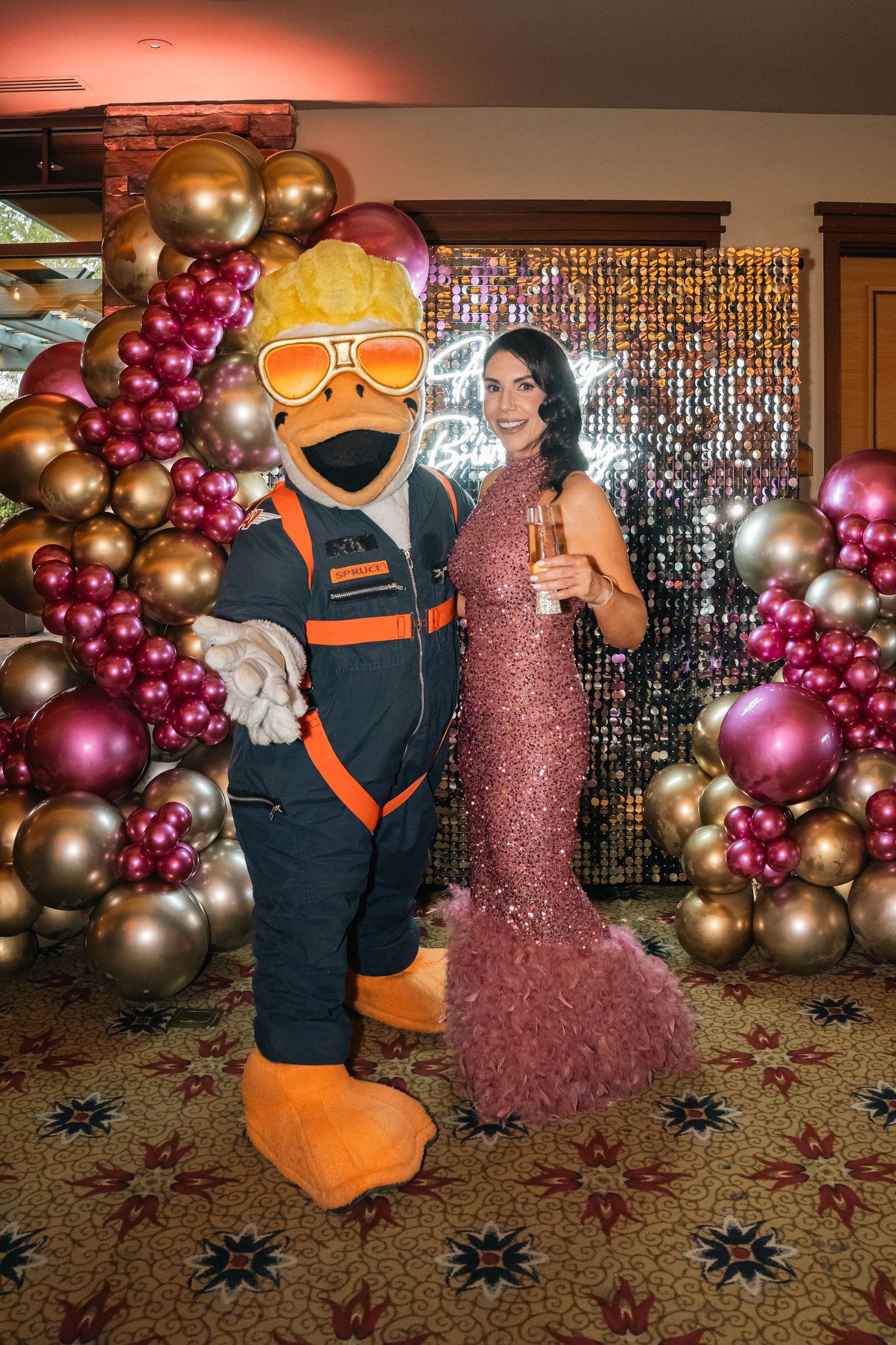 Person in mascot costume posing with someone in sparkly dress against balloon arch backdrop.