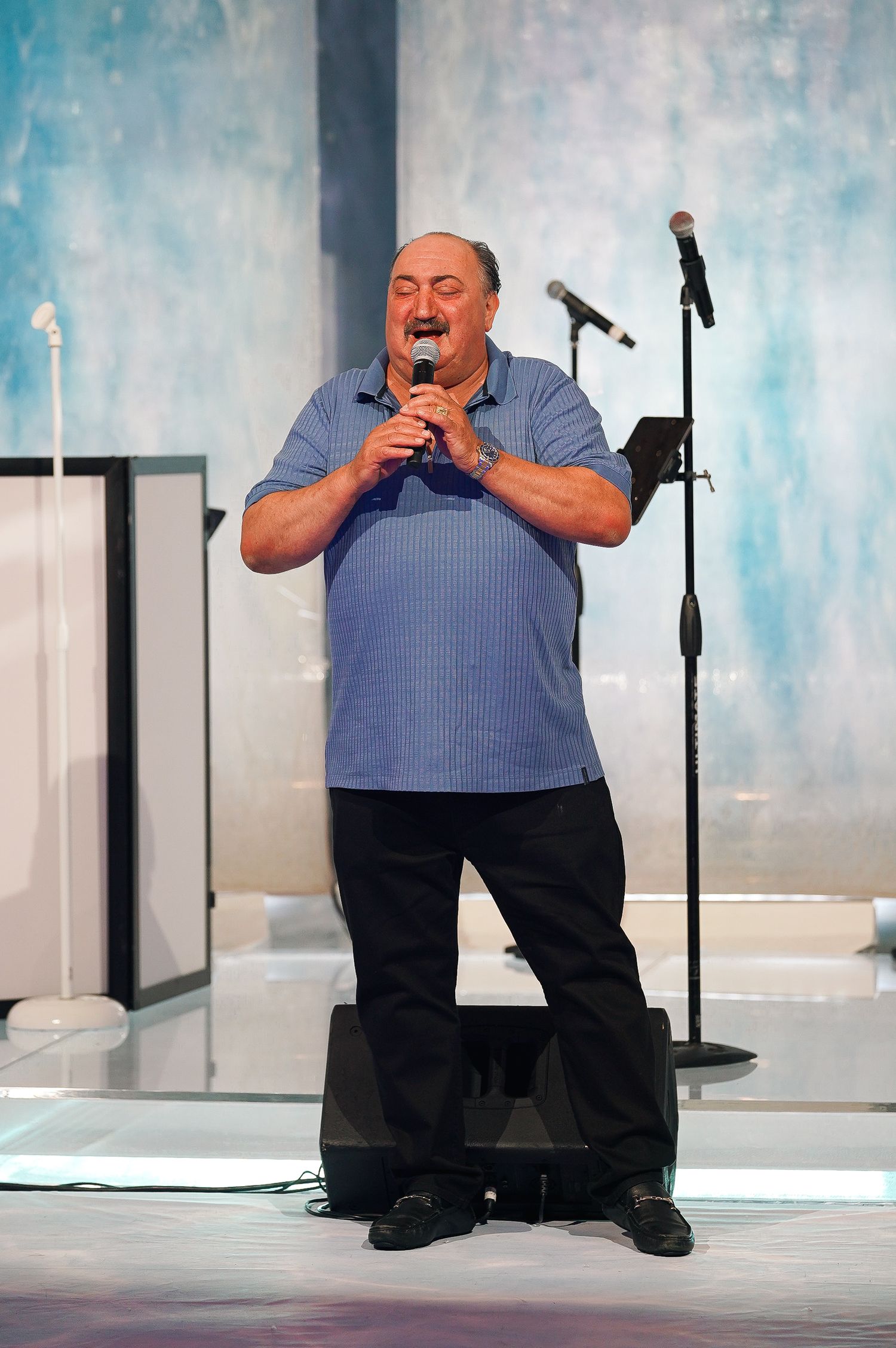 A performer in a blue shirt singing passionately on stage with microphone against a blue backdrop.