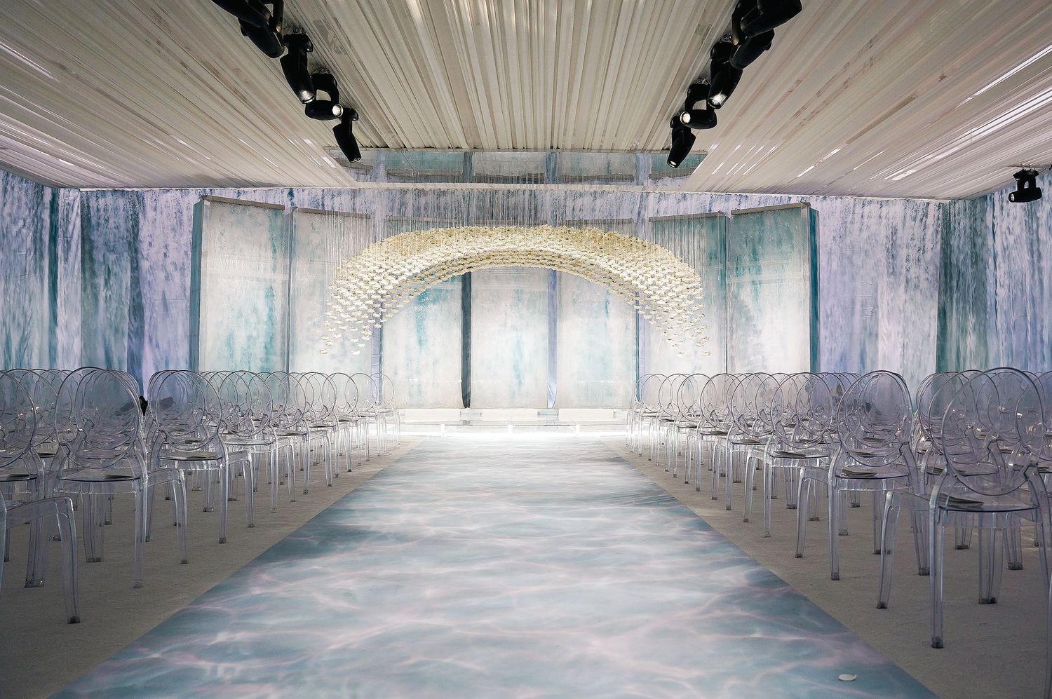 Wedding ceremony space with transparent chairs lined up facing illuminated floral arch.