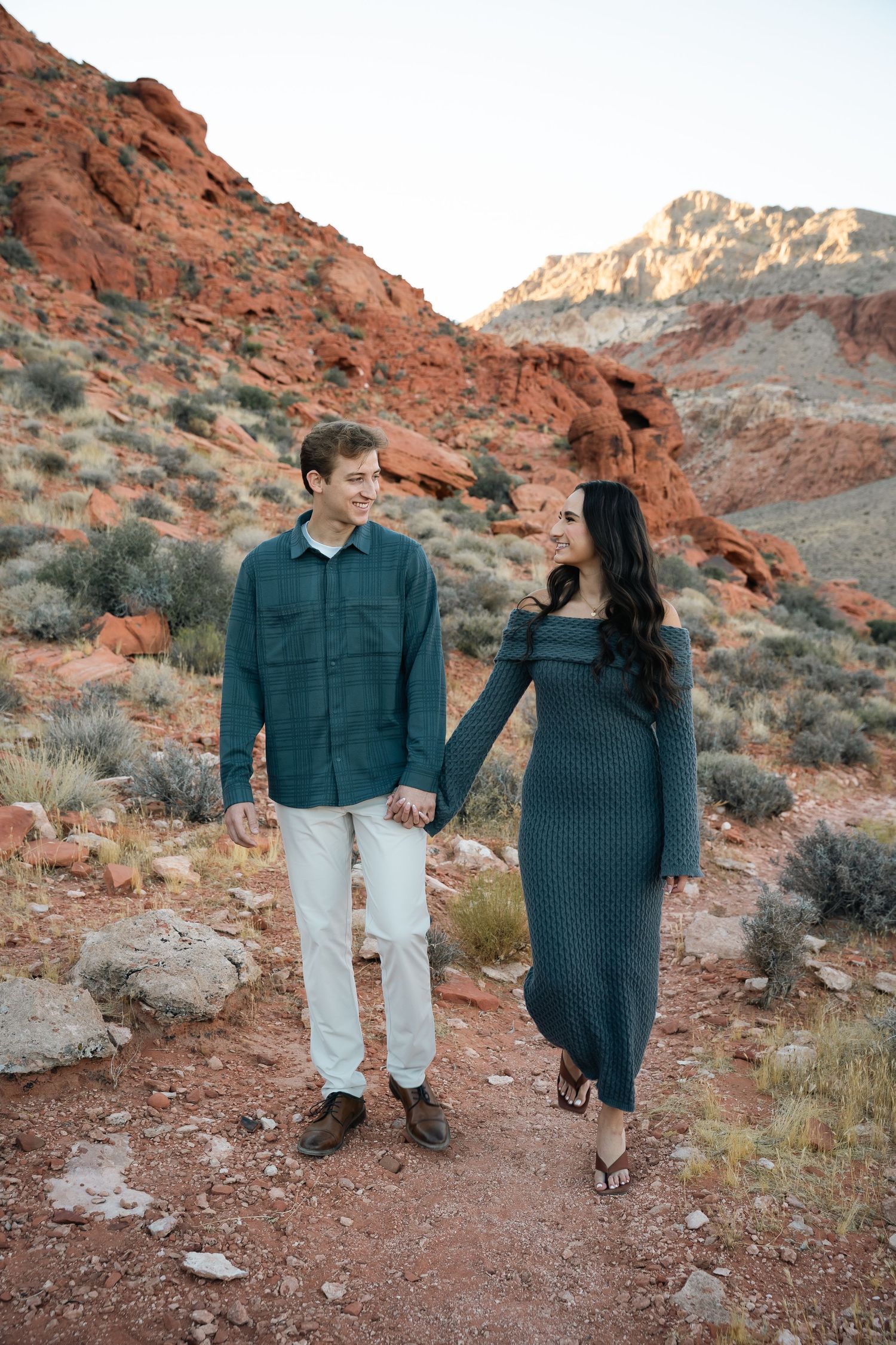 Couple holds hands while walking on desert trail surrounded by red rocks.