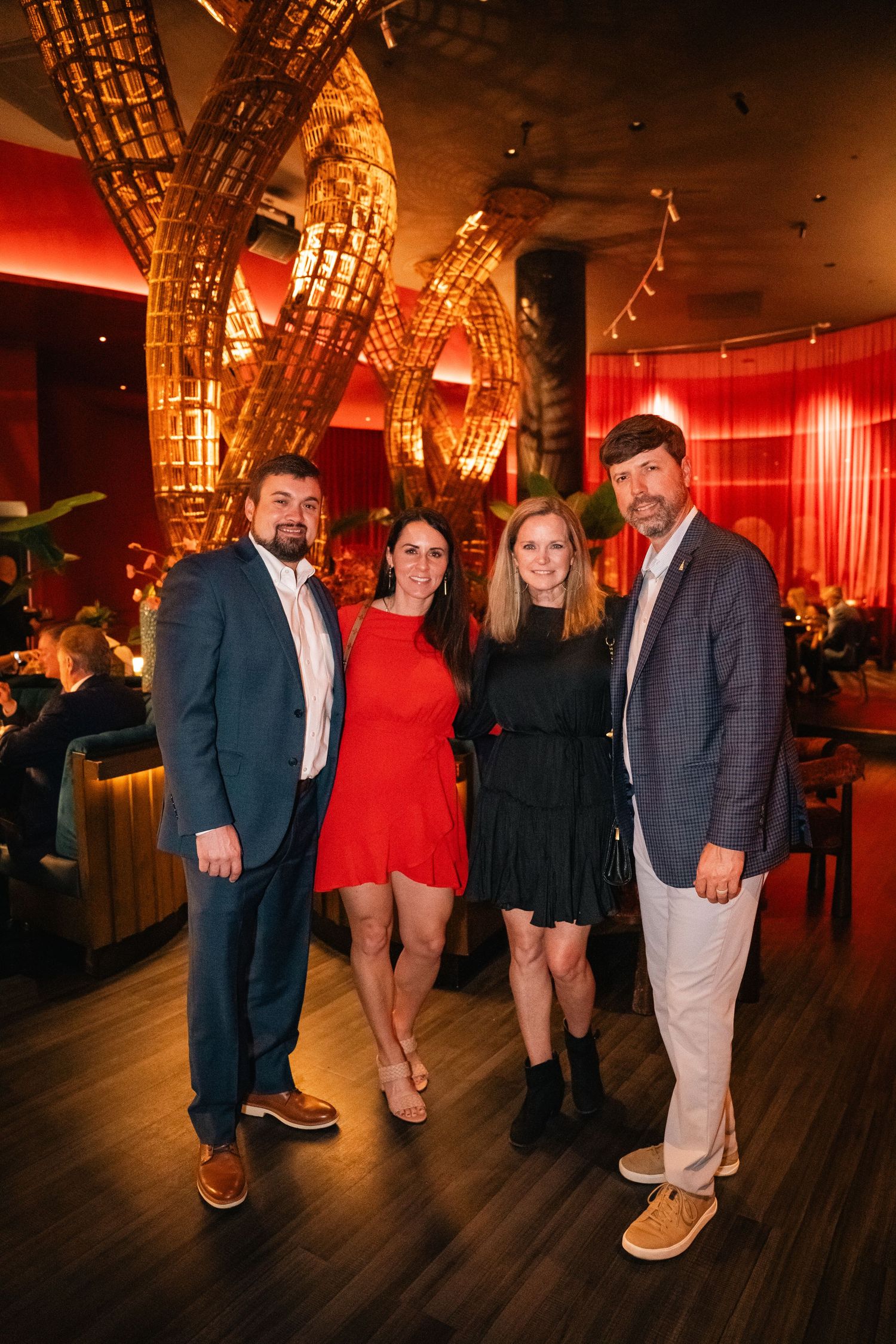 Two couples posing in formal attire at an upscale venue with dramatic lighting and wooden decor.