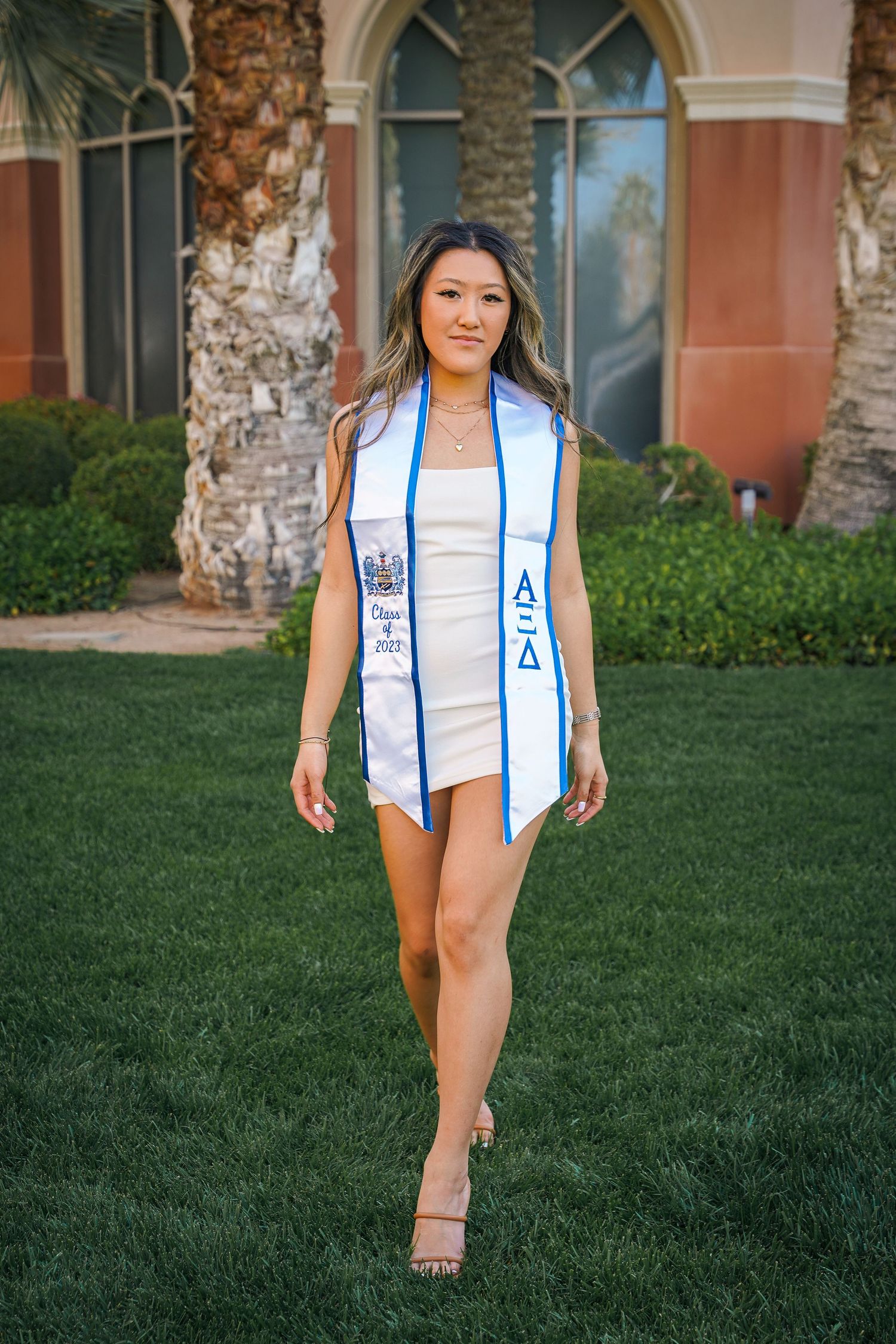 Graduate in white dress poses with graduation sash against Spanish colonial building and palm trees.