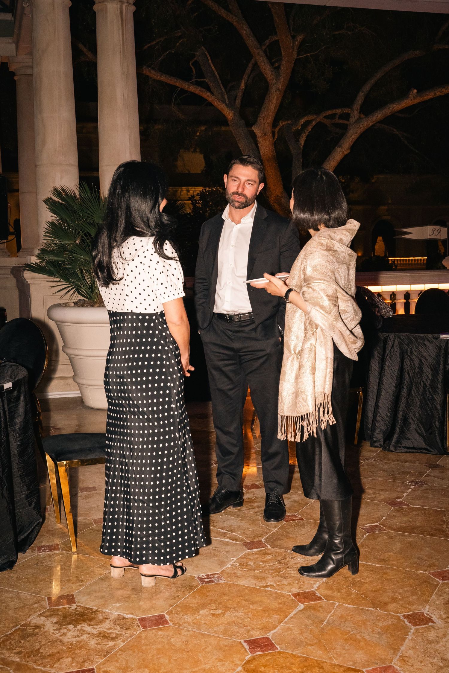 Party guests in formal wear interact on a marble floor near decorative planters.