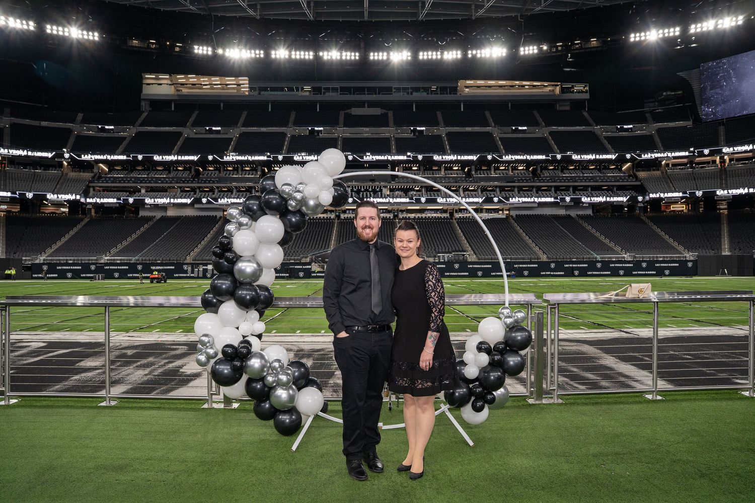 A couple poses on a football field with black and white balloon arch at a stadium venue.