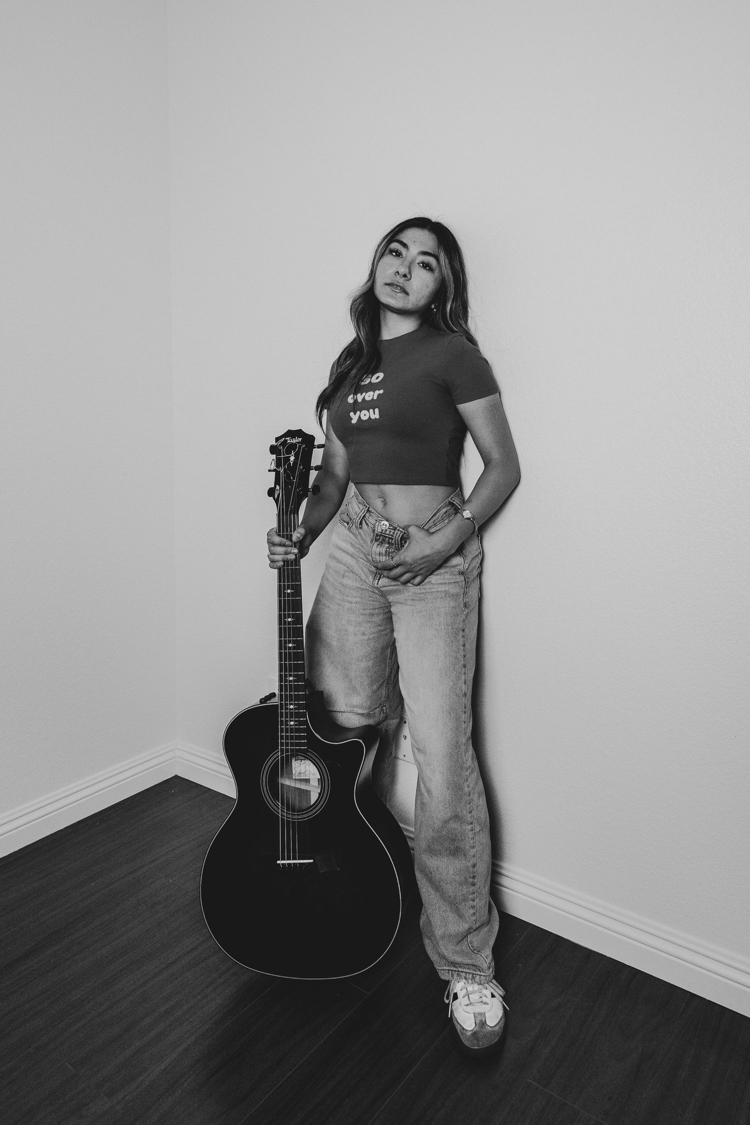 Black and white photo of someone in casual wear posing with an acoustic guitar against a plain wall.