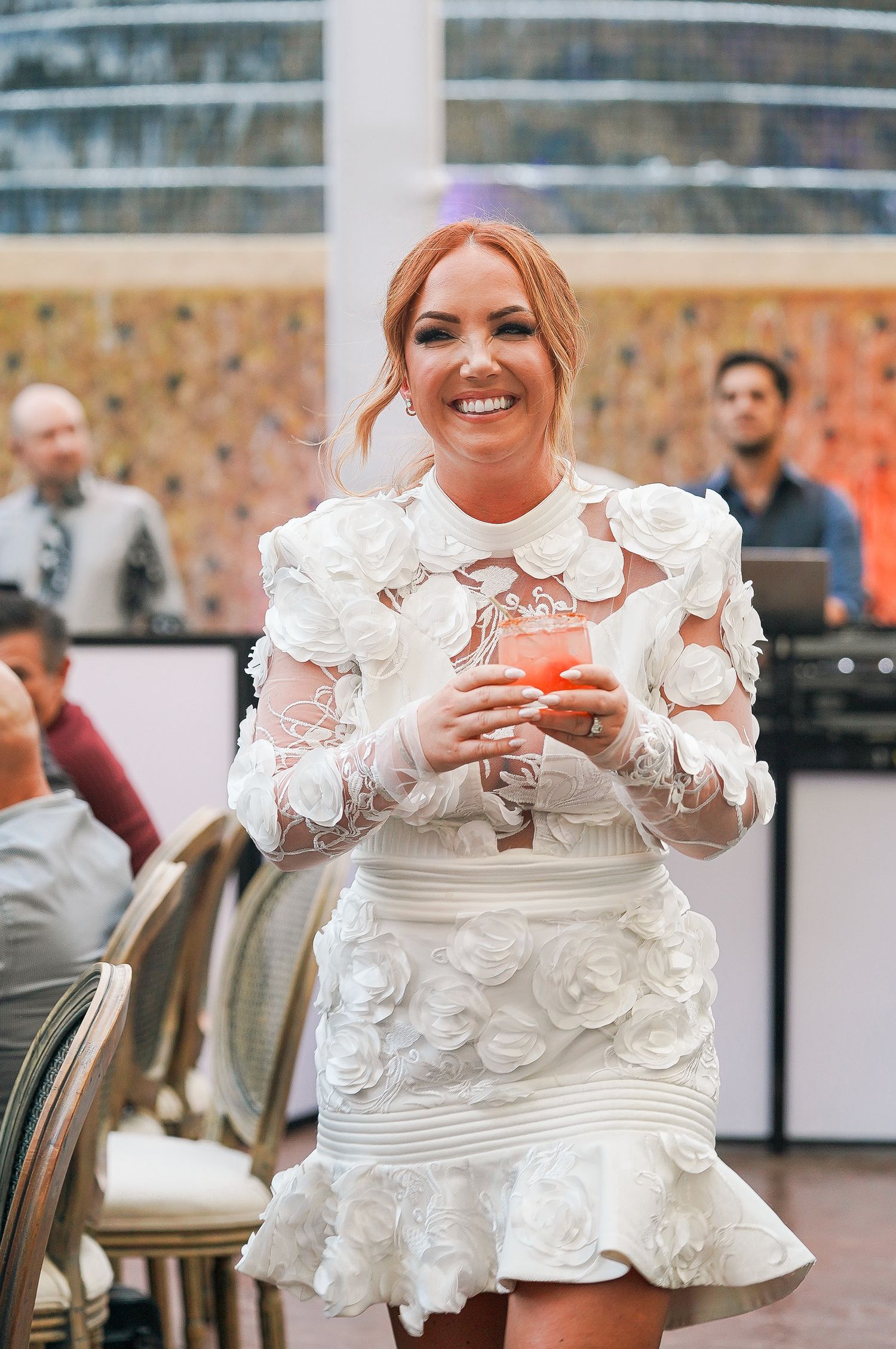 Person in white textured dress smiles while holding cocktail at outdoor event.
