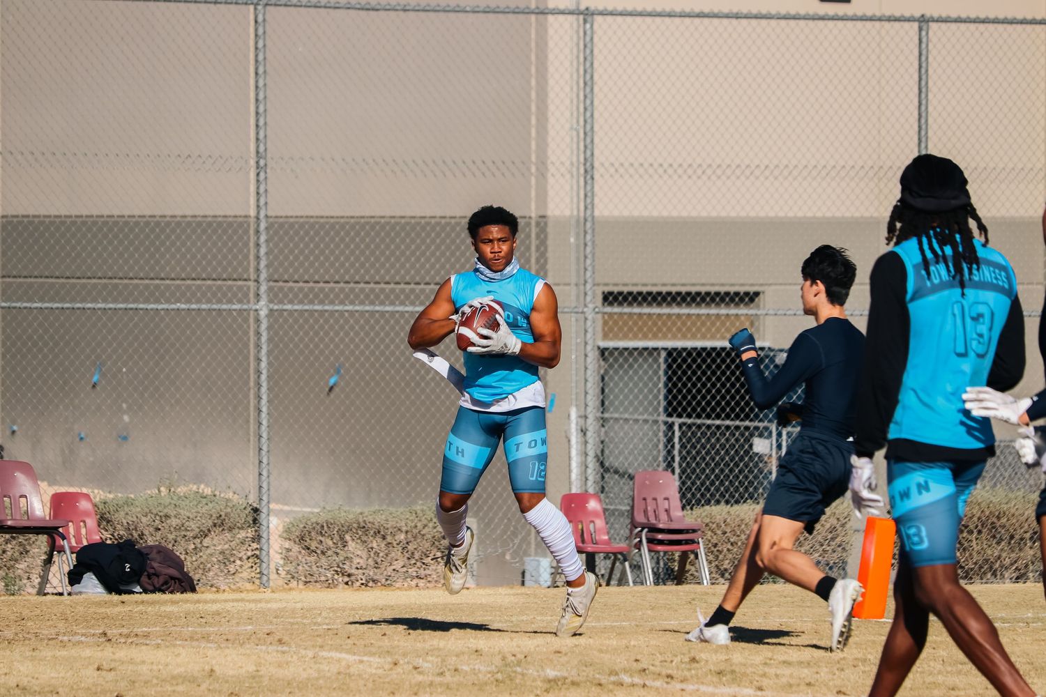 Athletes play flag football on a sunny field next to a chain-link fence.