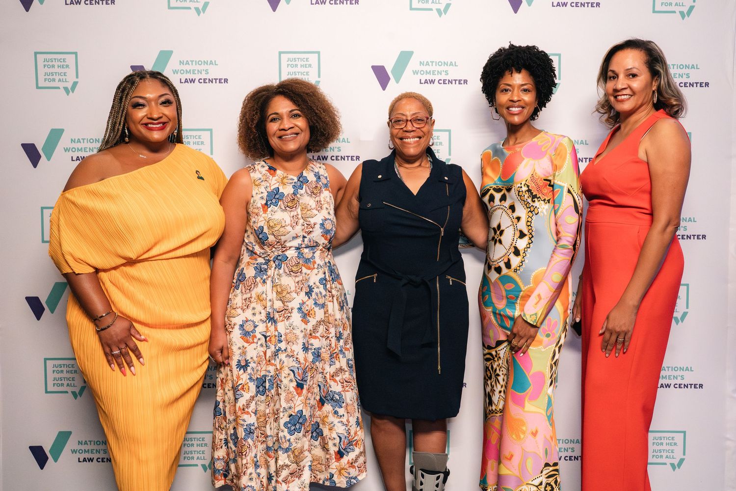 Group of women in colorful dresses posing at formal event with branded step-and-repeat background.