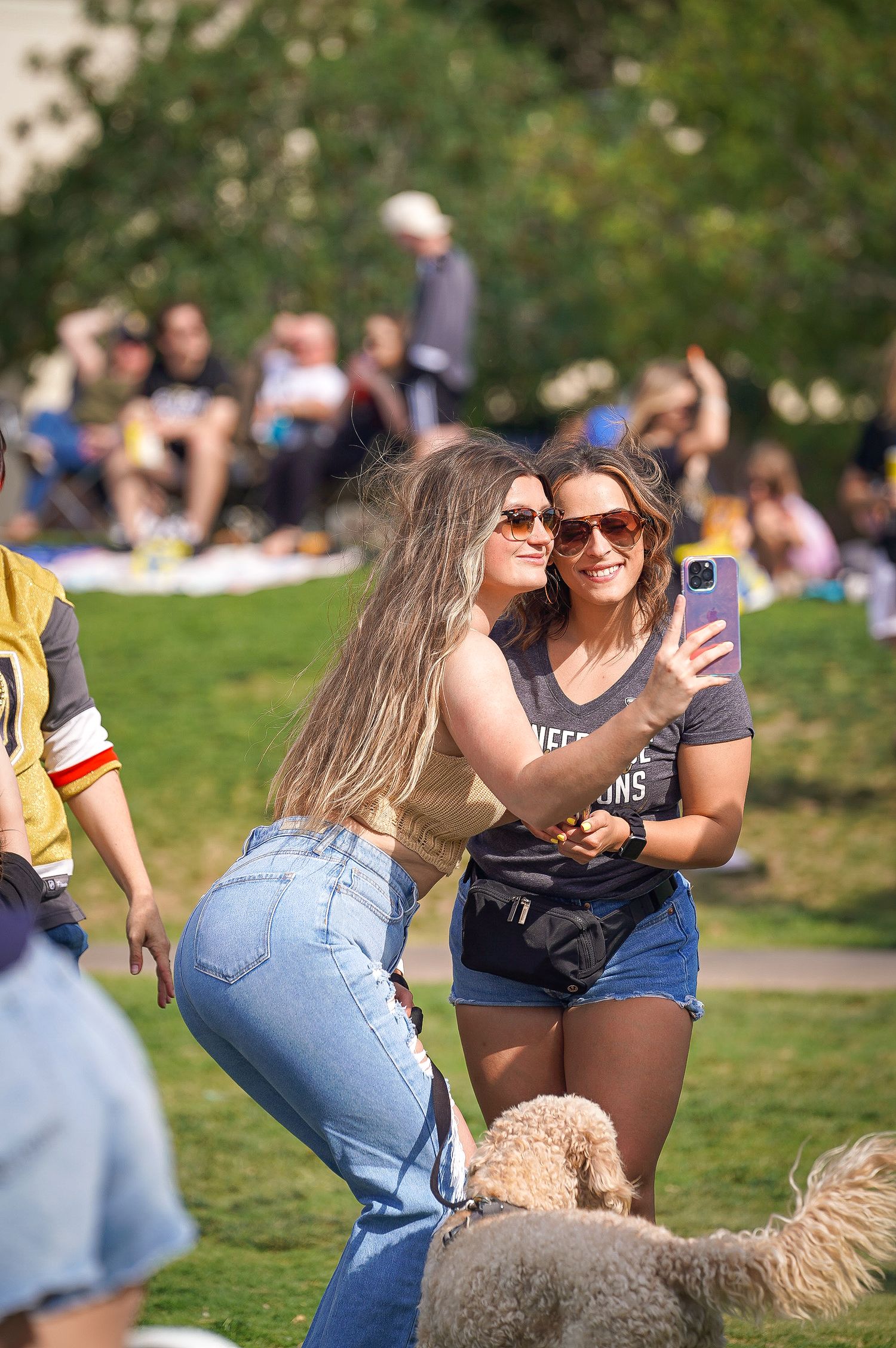Two friends taking selfies together on grassy lawn at outdoor festival.