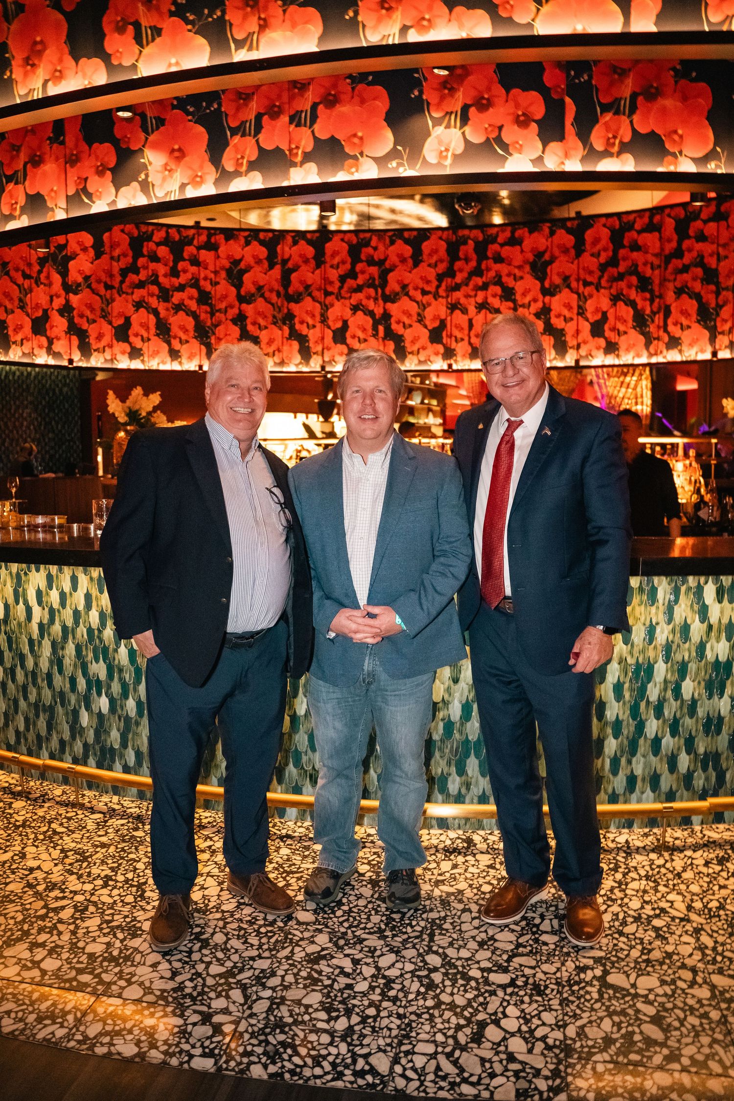 Three men pose together in front of a decorative red and orange illuminated wall panel.