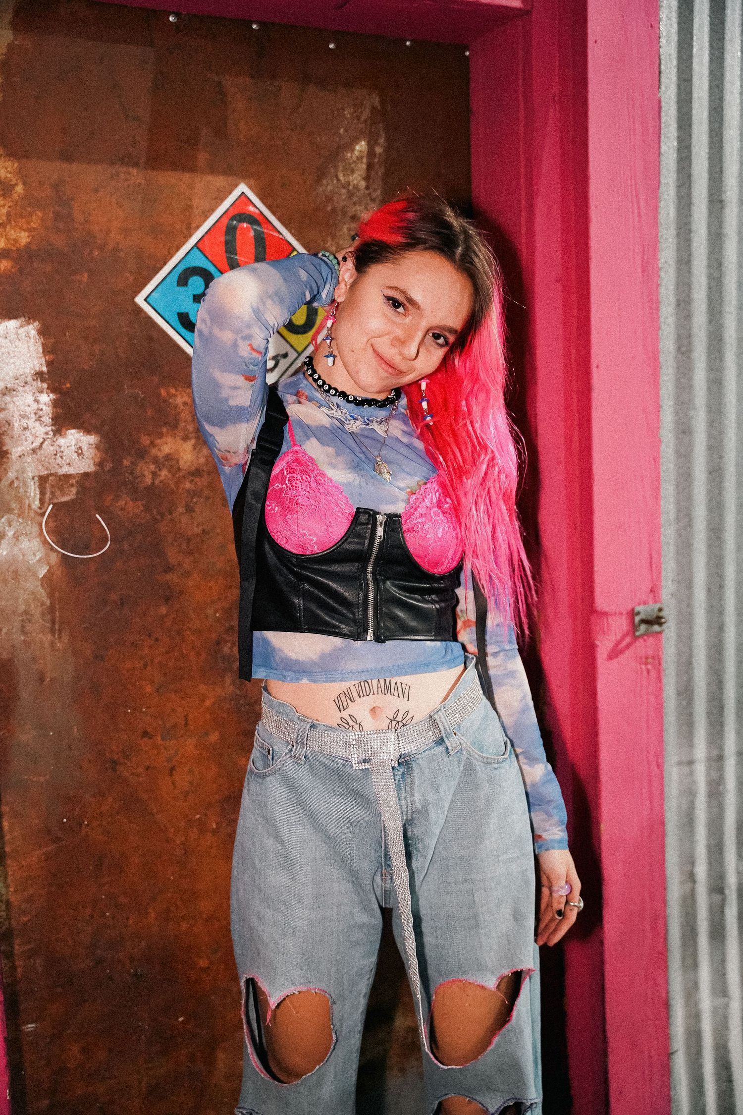 Model with pink hair poses in distressed denim against pink and rusty metal backdrop.