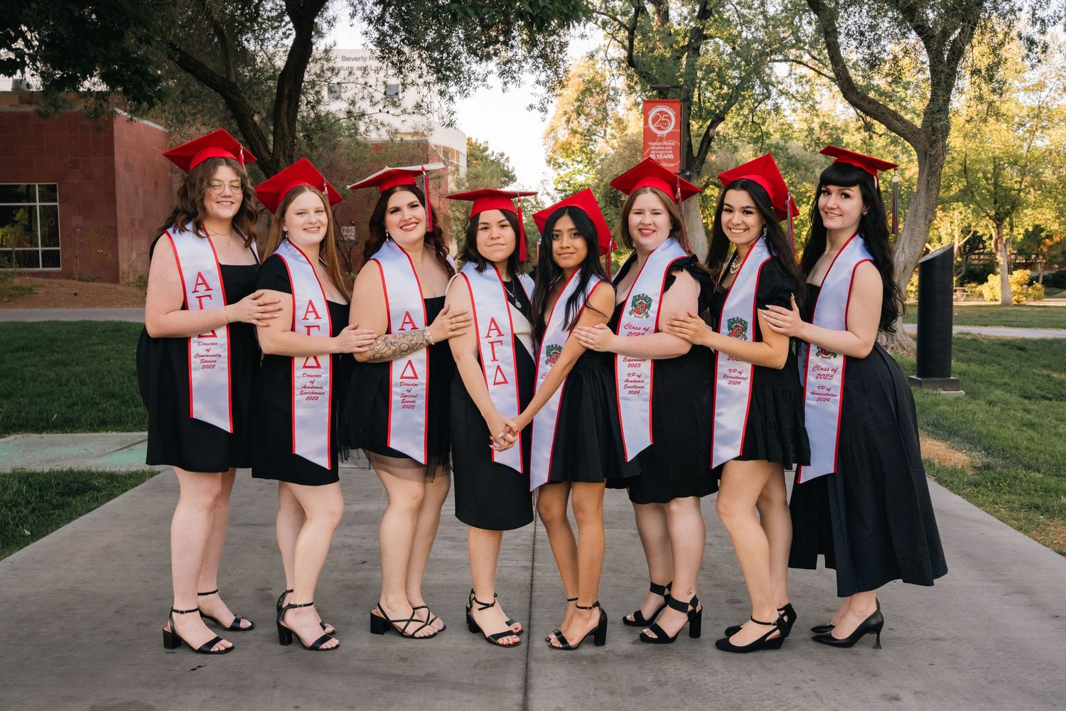 Graduates in black dresses and pink sashes pose together wearing red graduation caps.
