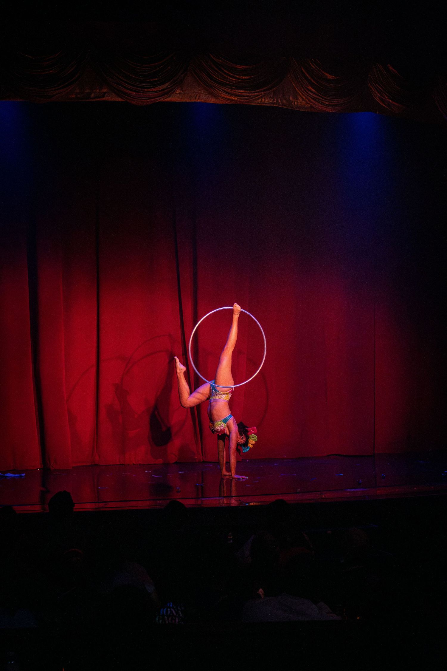 Aerial hoop artist performing against red stage curtain under blue spotlights.