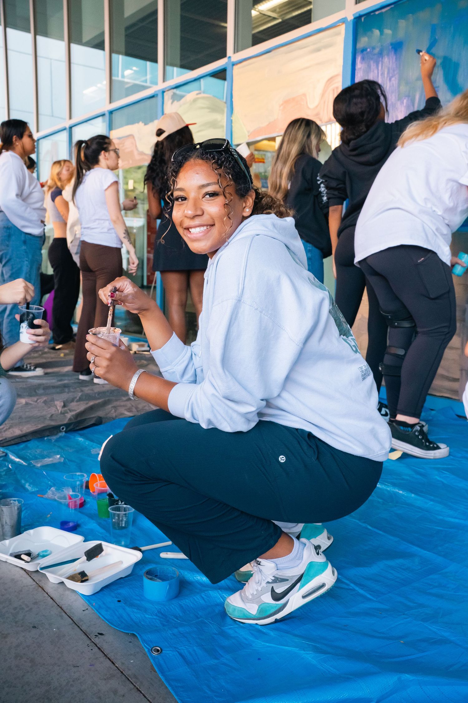 Students participate in an outdoor art project on a blue tarp while painting colorful murals on a wall.