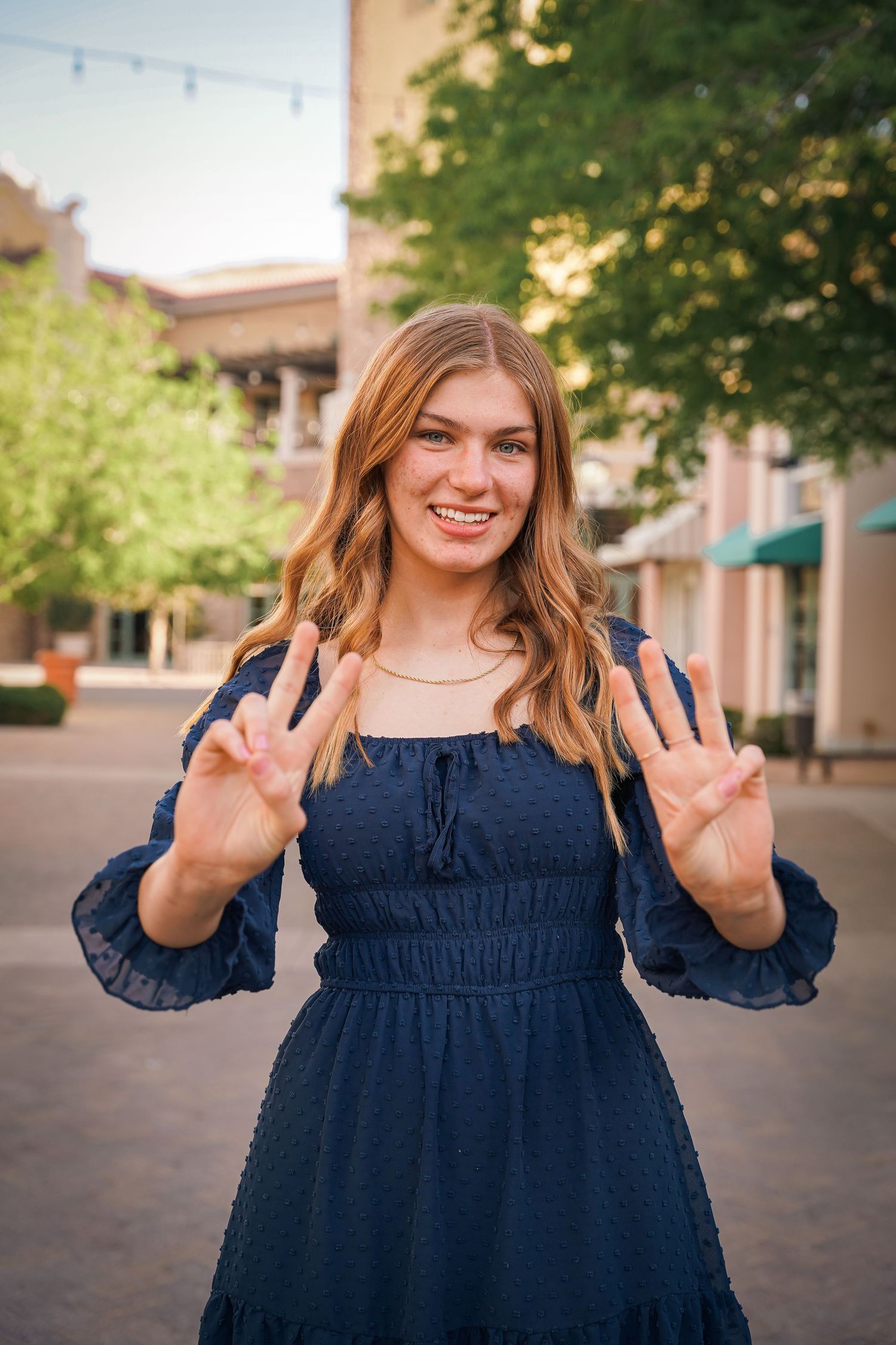 Person in navy dress making peace signs while standing on outdoor shopping plaza.