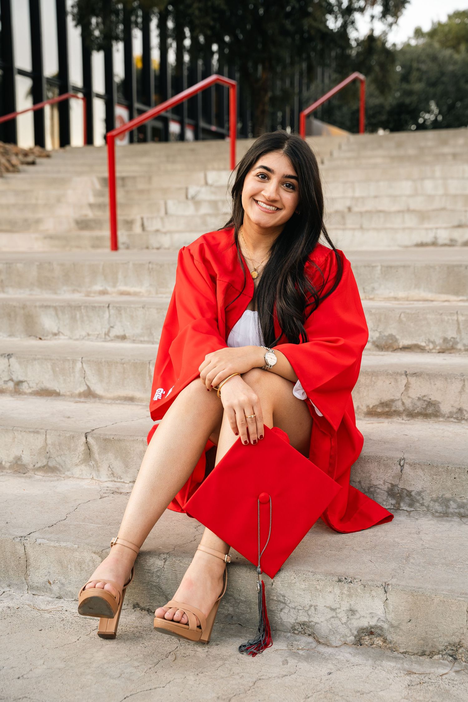 Graduate in red graduation gown sitting on concrete steps outdoors.