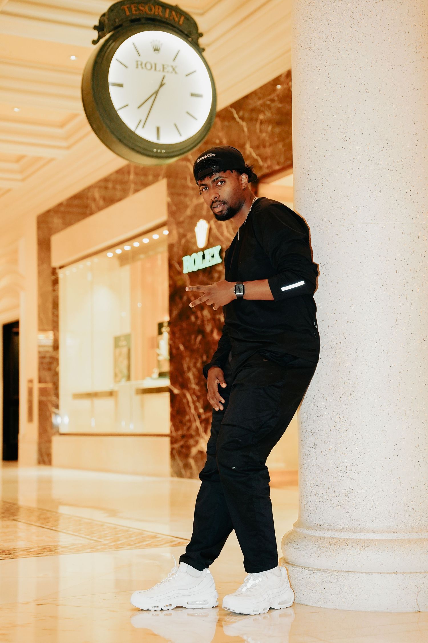 Person in all black outfit standing beneath large Rolex clock in luxury interior.