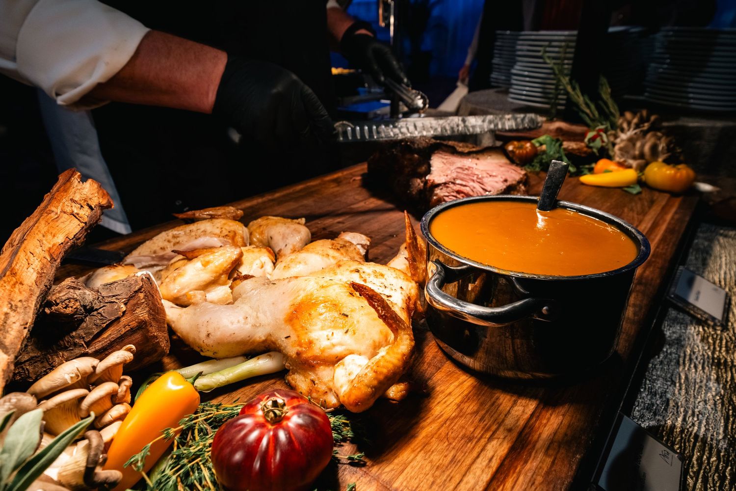 A rustic wooden table displays roasted poultry, vegetables, and a pot of orange soup in warm lighting.
