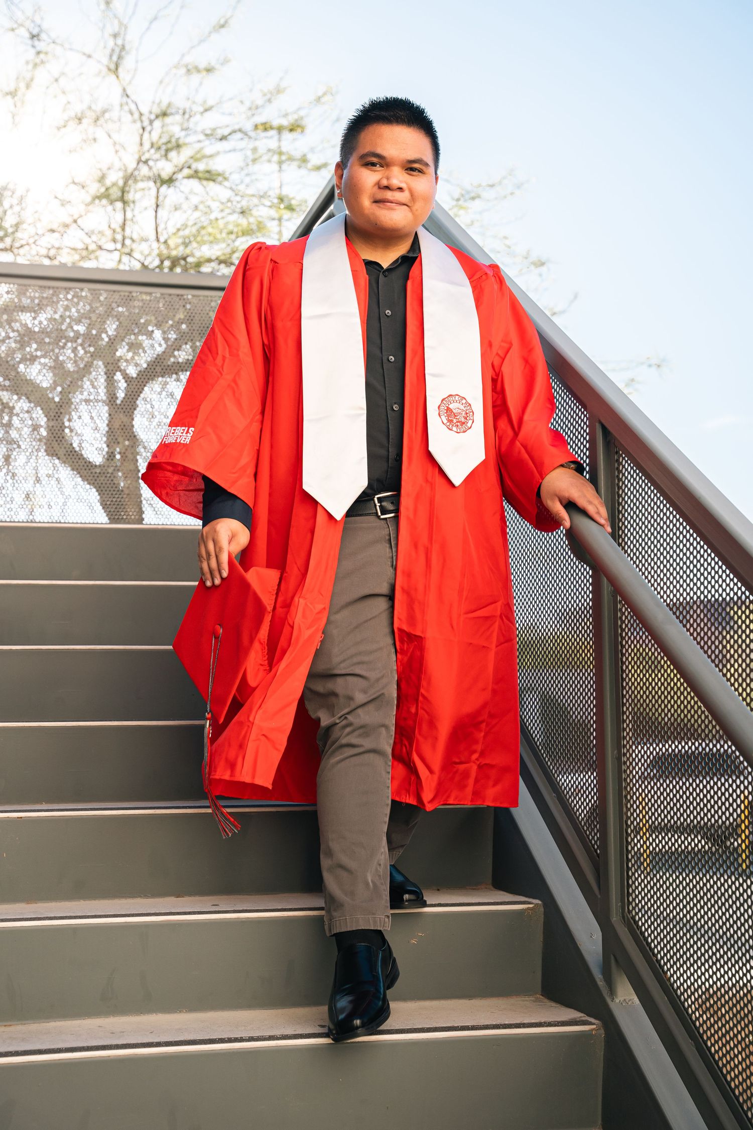 Graduate in red academic regalia descends outdoor metal staircase at university campus.