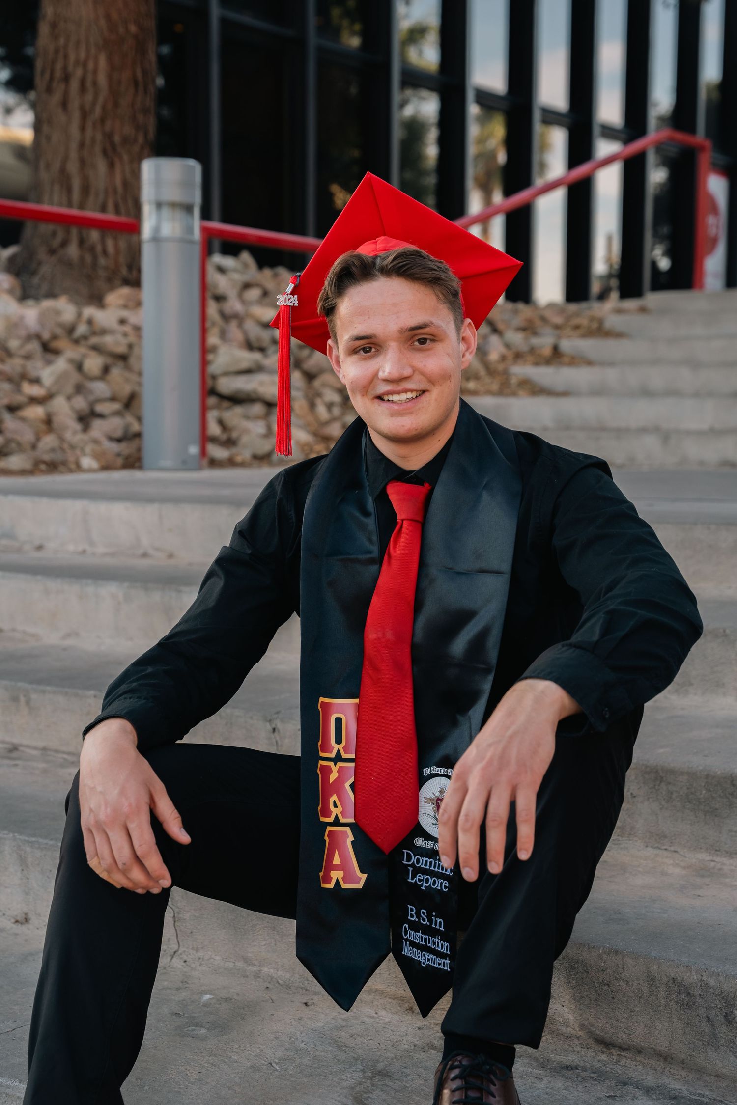 Graduate in cap and gown sits on steps wearing red tie and DXA graduation stole.