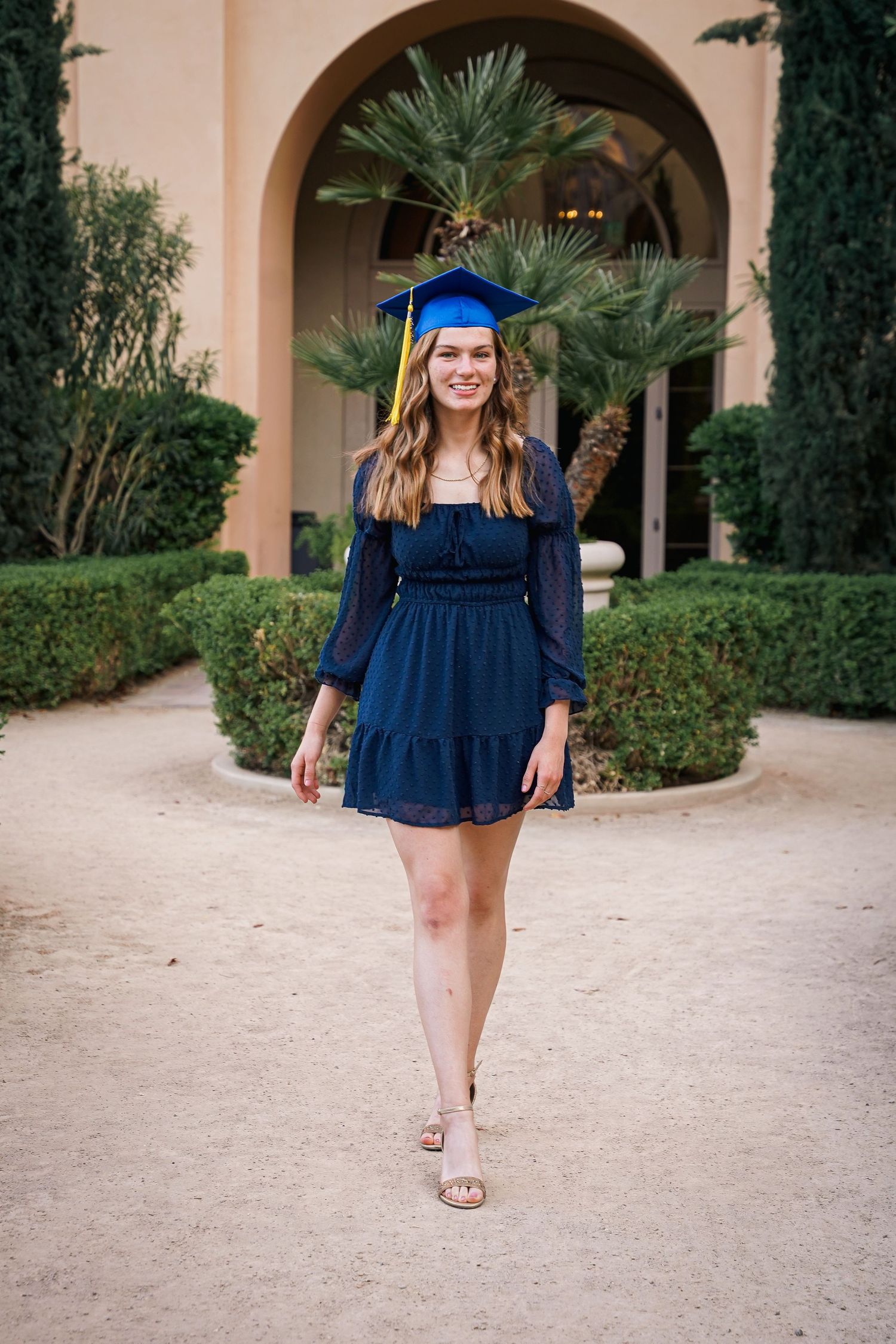 Graduate in blue dress and cap poses on Mediterranean-style campus path.