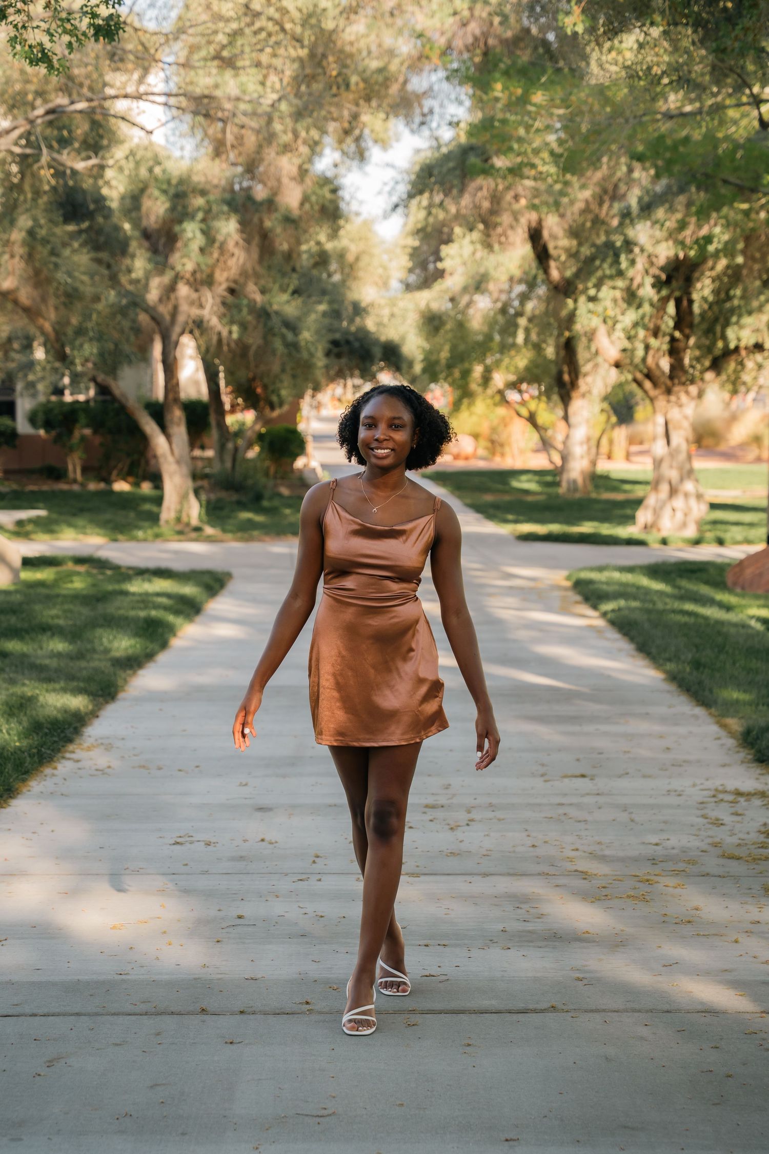 Woman in brown dress walking confidently along tree-lined pathway in a sunny park setting.