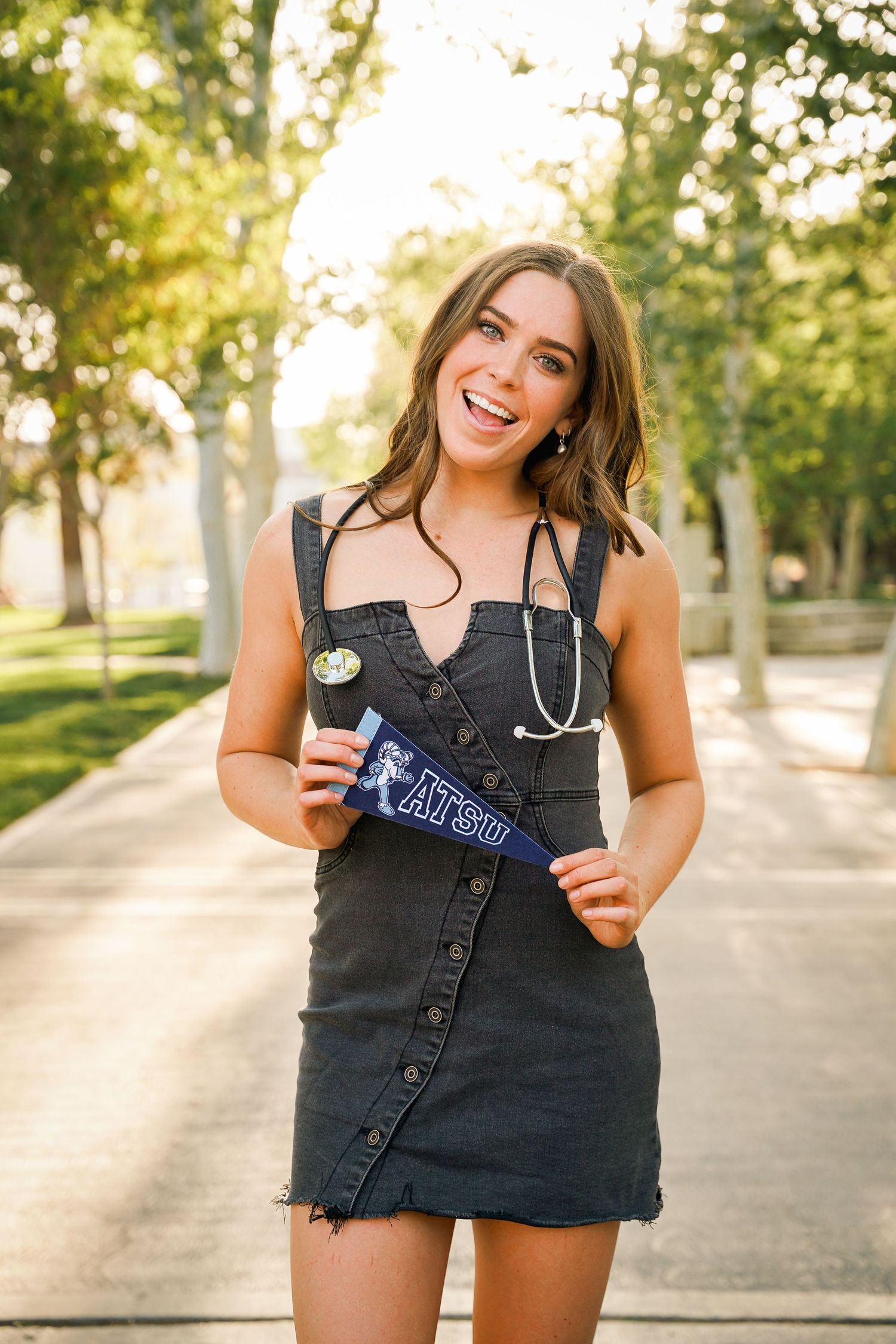Graduate in black denim dress celebrating with stethoscope and university memorabilia in sunny outdoor setting.