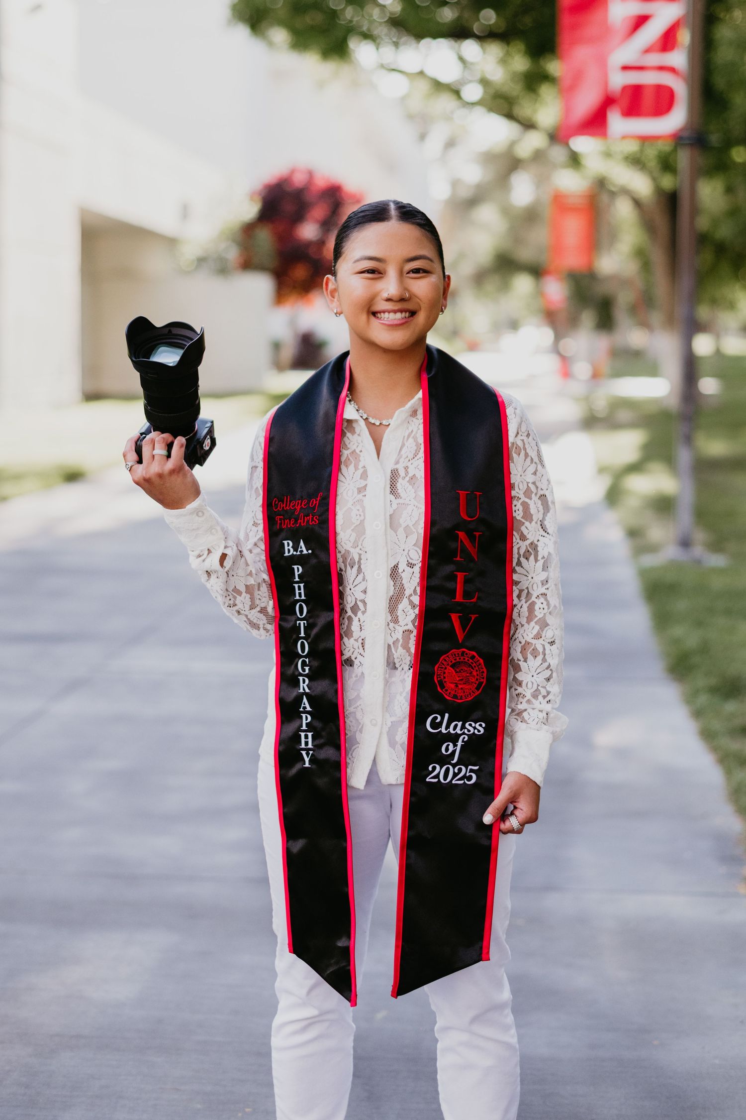 JJ Monaco. Graduate in white lace top and graduation stole holding camera outdoors. Monaco Media.