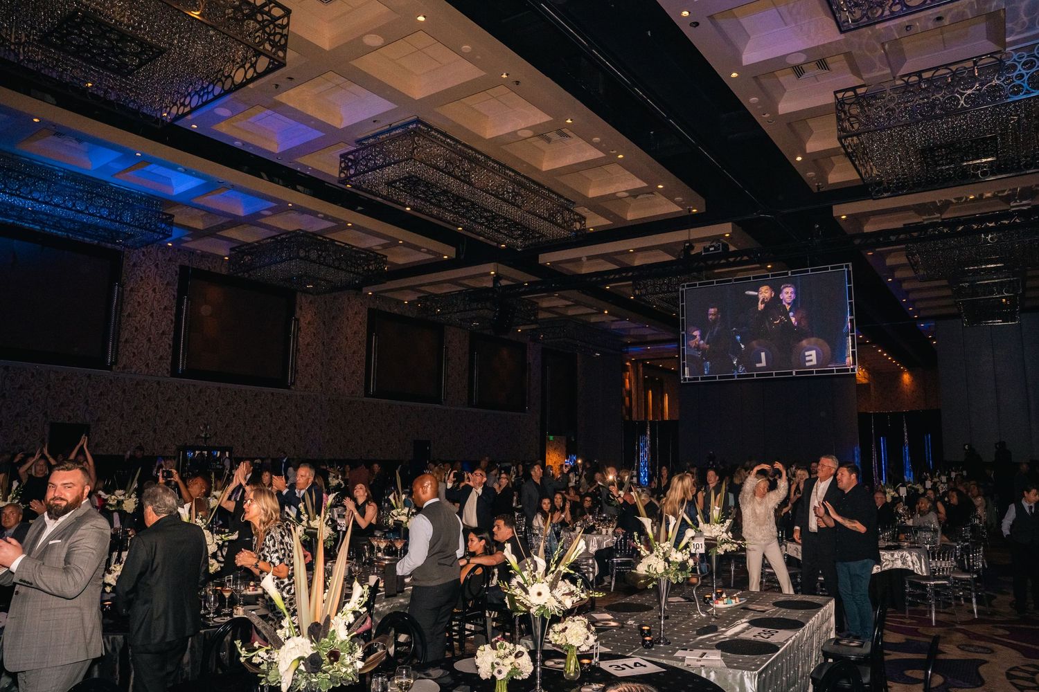 Large crowd stands and cheers in an elegant ballroom with blue lighting and crystal chandeliers.