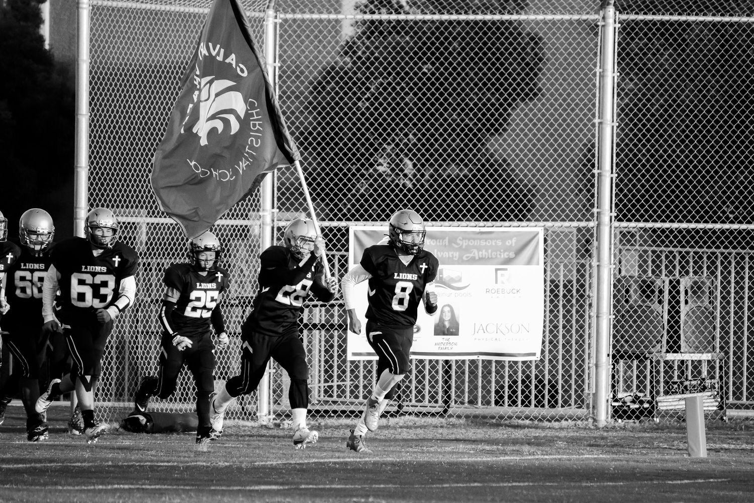 Team runs onto field carrying flag during black and white sports photography.