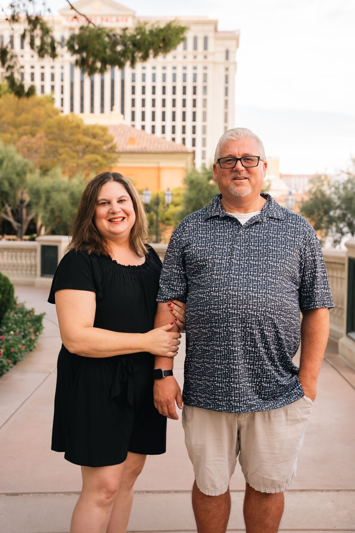 Couple poses together on outdoor walkway with Las Vegas hotel architecture in background.