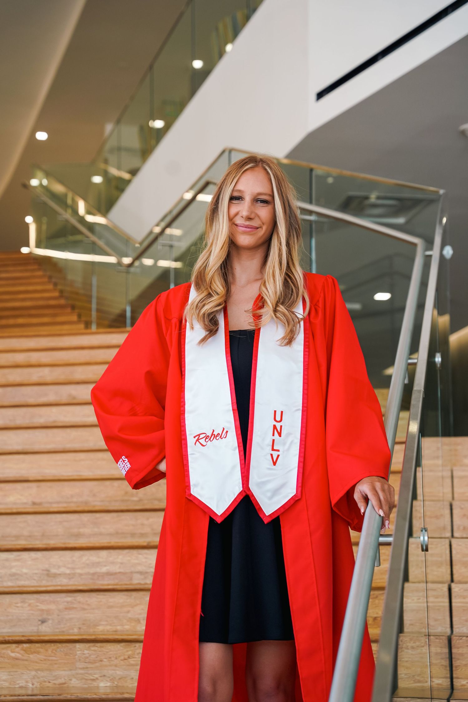 Graduate in red gown and white stole poses on modern staircase indoors.