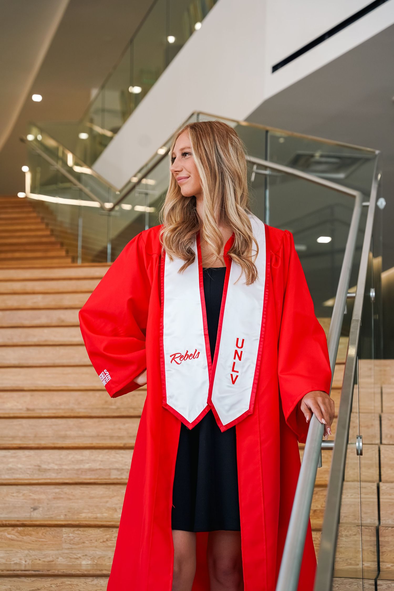 Graduate in red graduation gown and white stole poses on wooden staircase in modern building.