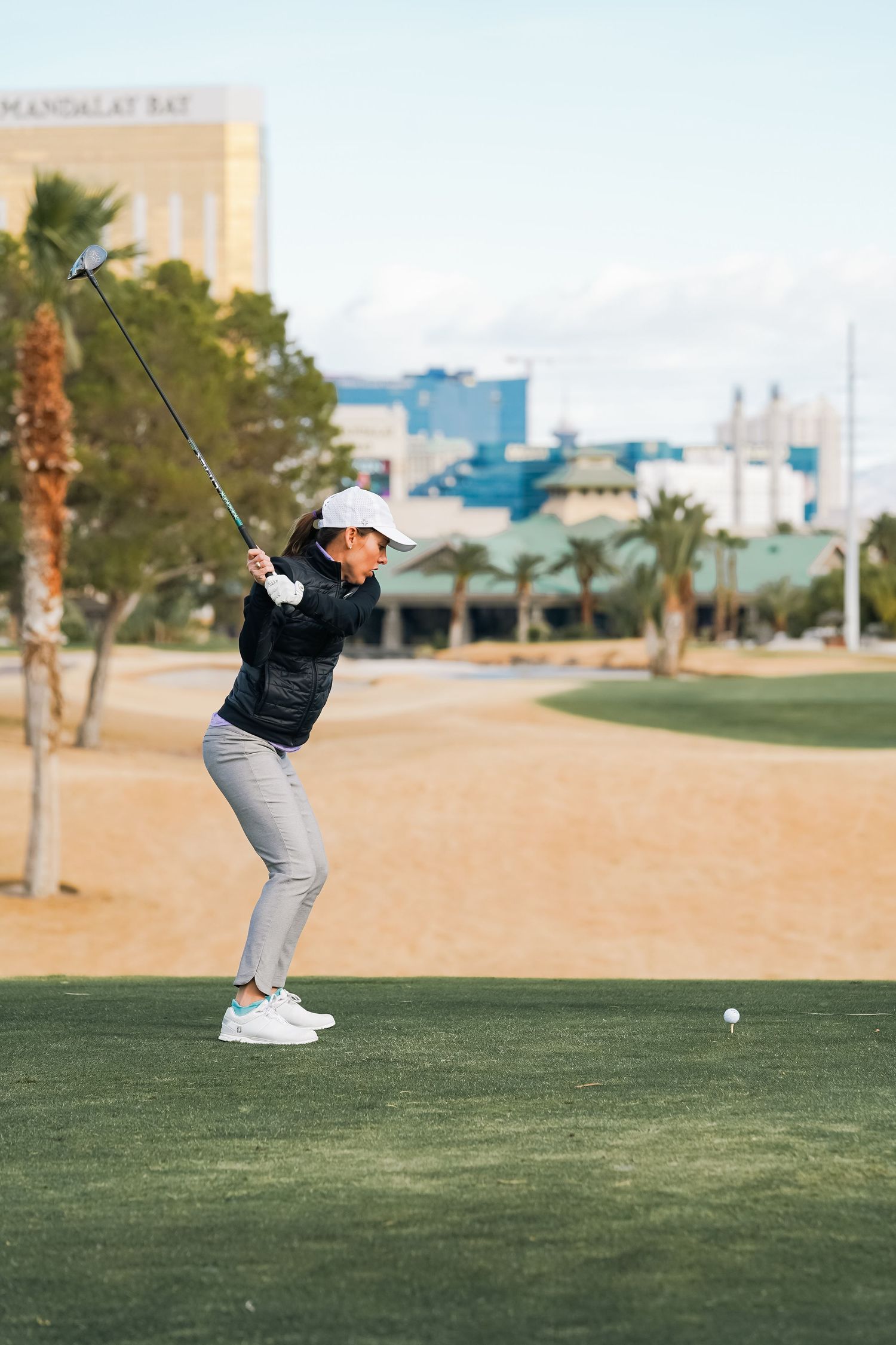 Golfer practicing swing form on desert golf course with palm trees and city skyline in background.
