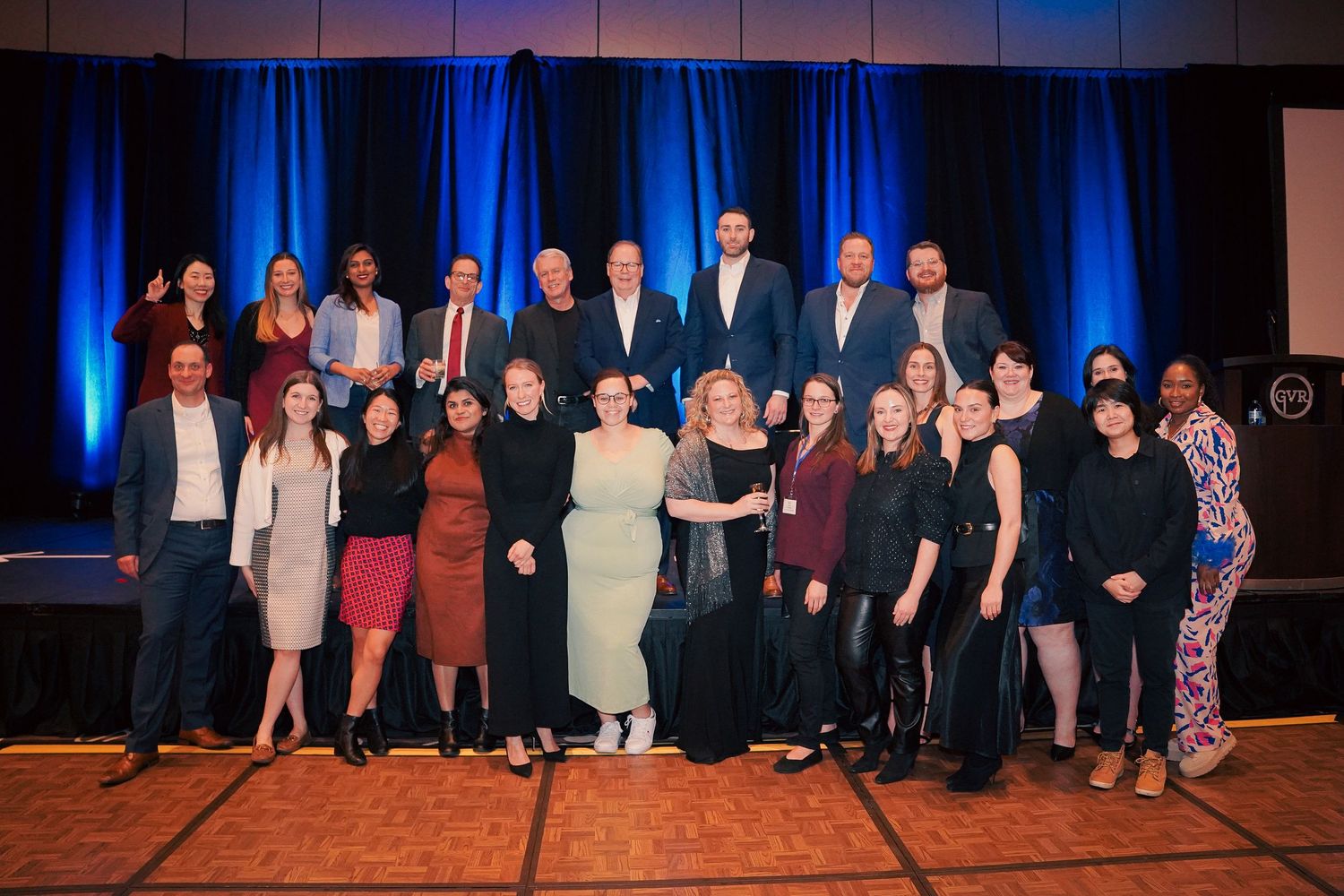 Group photo of professionally dressed people posing together on stage with blue backdrop.