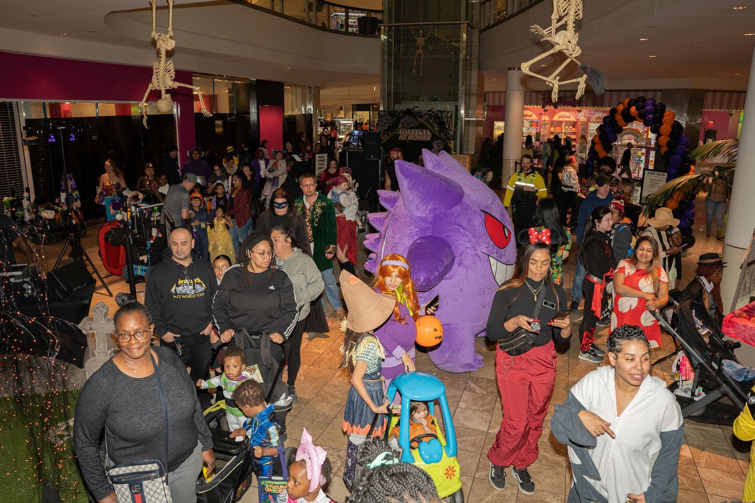 Crowd of Halloween party attendees gathered beneath decorative purple and orange balloons.