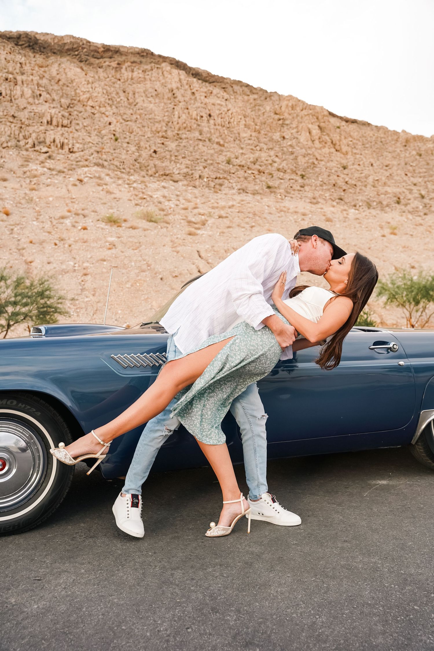 Romantic dip pose by vintage blue car with desert mountains in background.