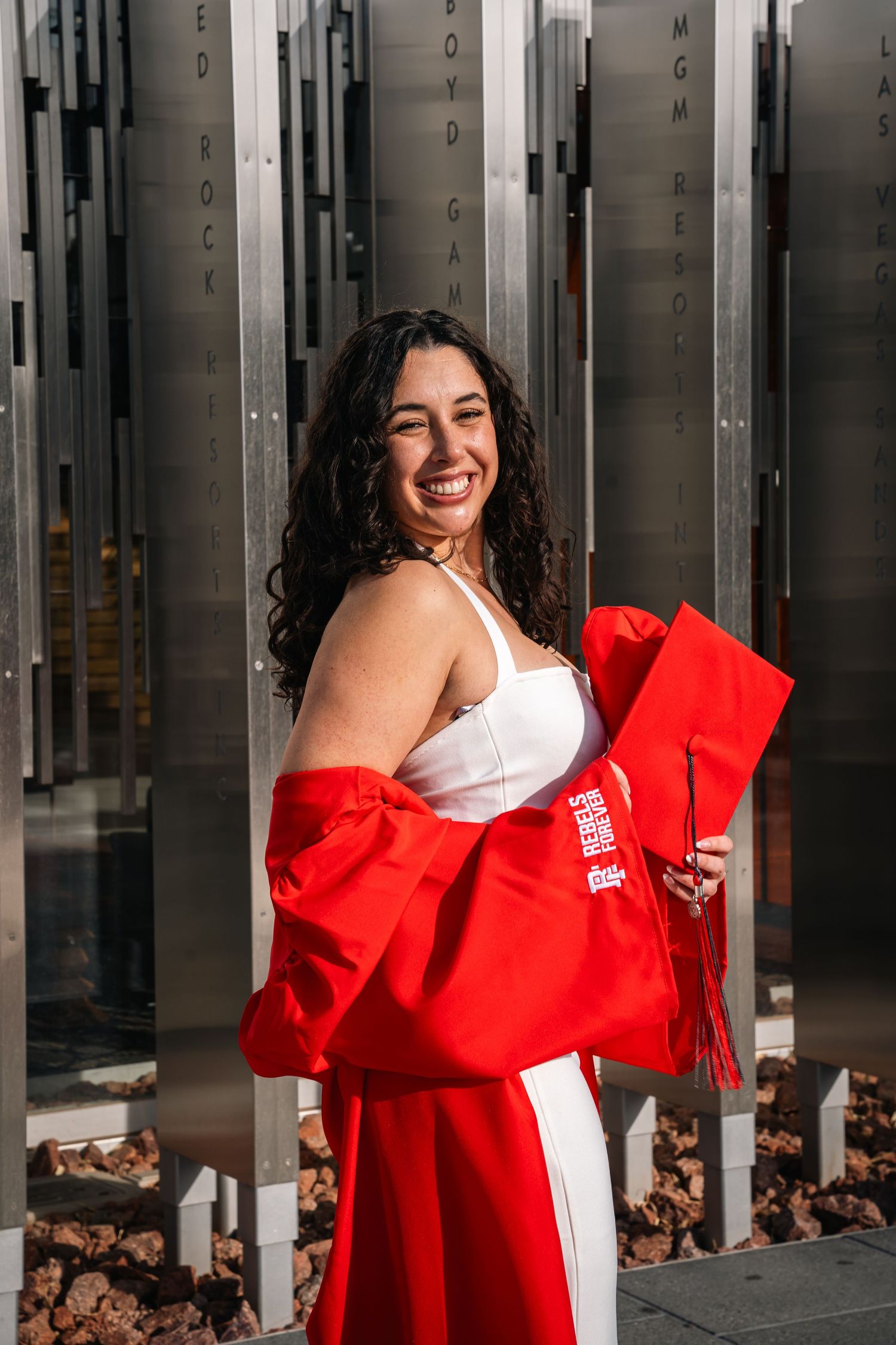Graduate in white dress and red graduation gown poses by modern metal architecture.