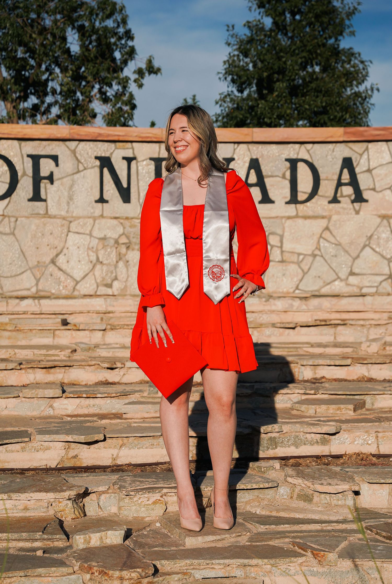 Graduate in red graduation regalia poses against stone wall with University of Nevada sign.