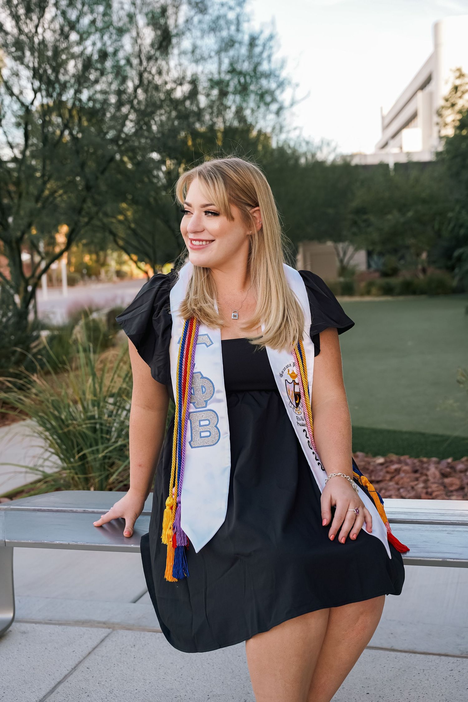 Graduate in black dress with decorated stoles poses outdoors against landscaped campus background.