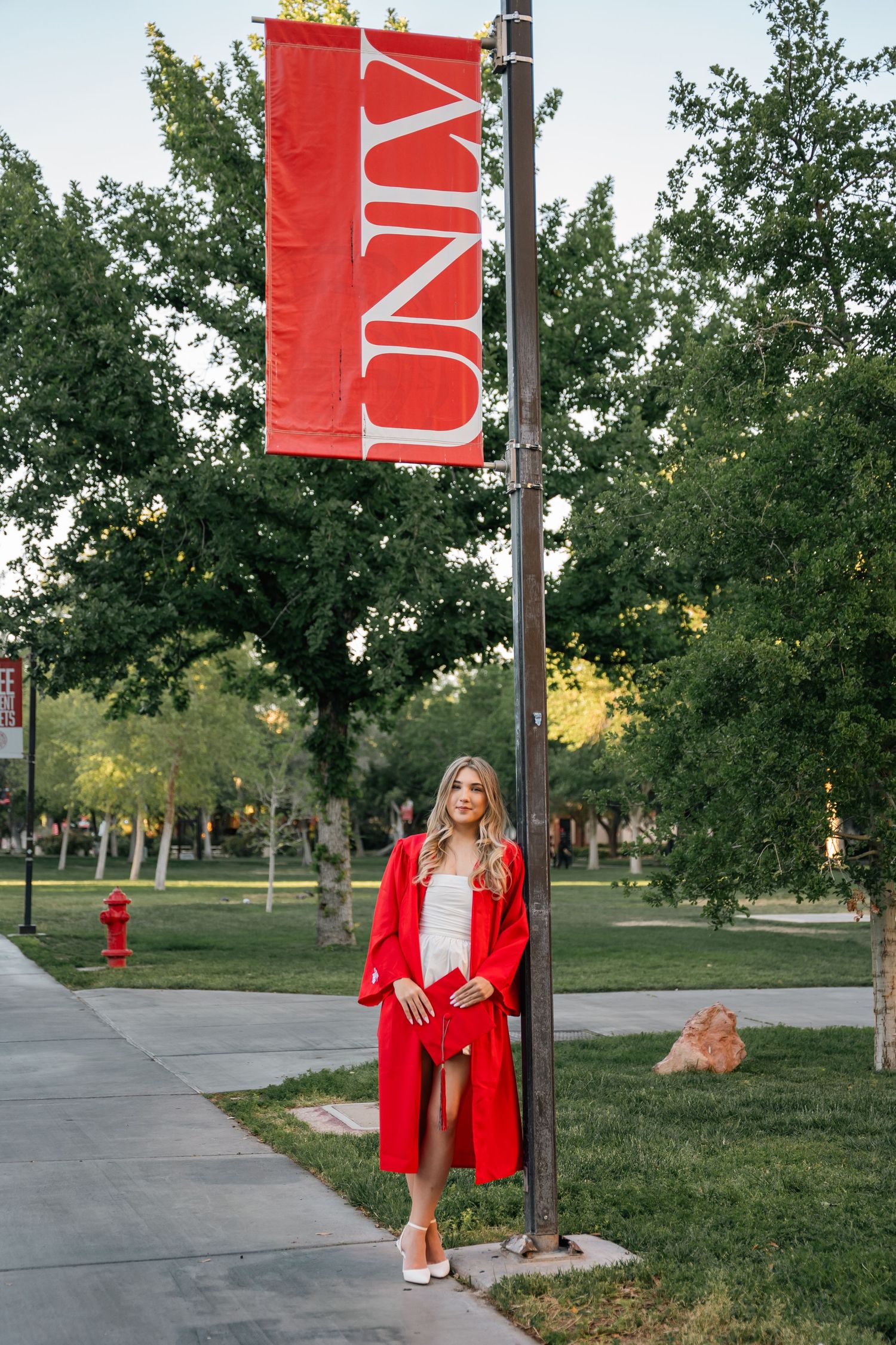 Graduate in red cap and gown poses by UNLV banners outdoors on campus walkway.