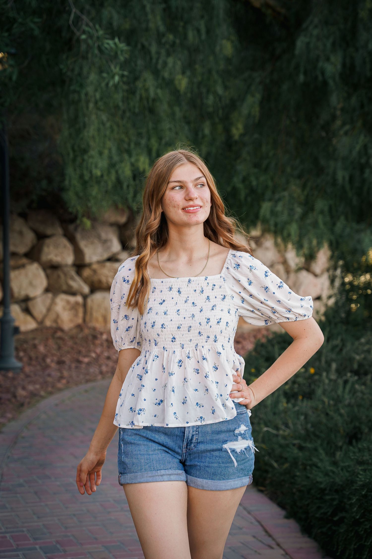 Young woman in floral puff sleeve top poses outdoors near stone wall.