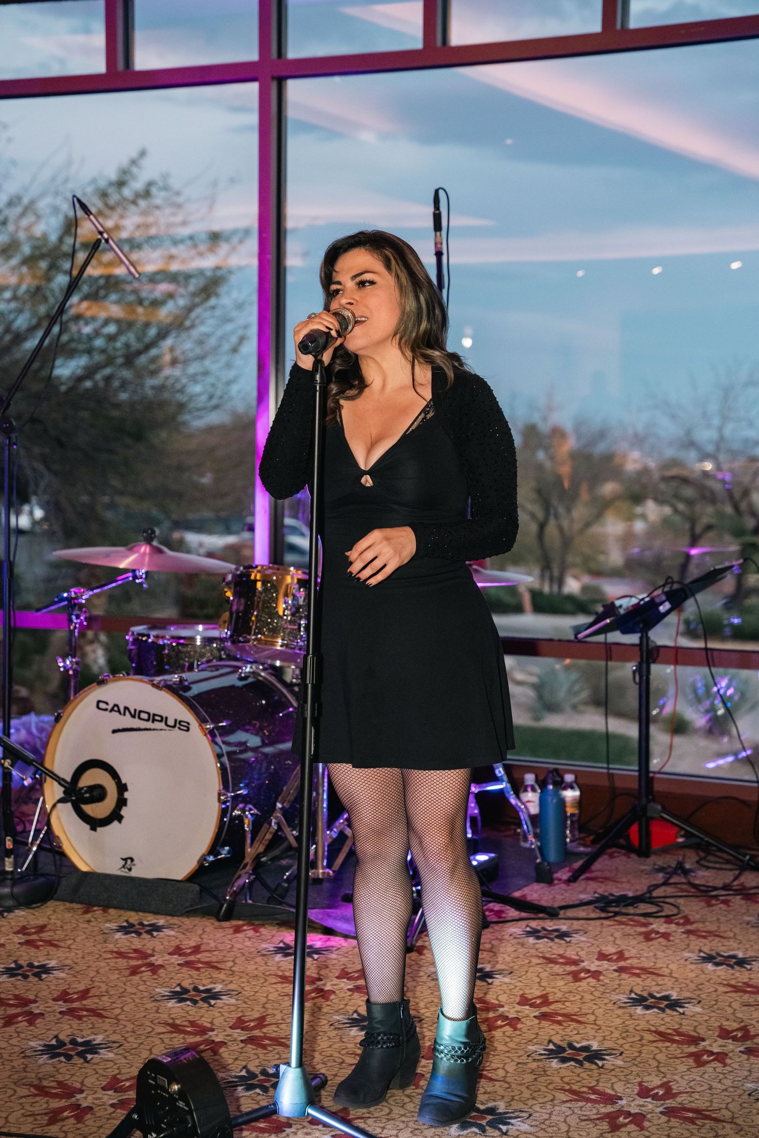 Singer performing with microphone in black dress at venue with drum set.